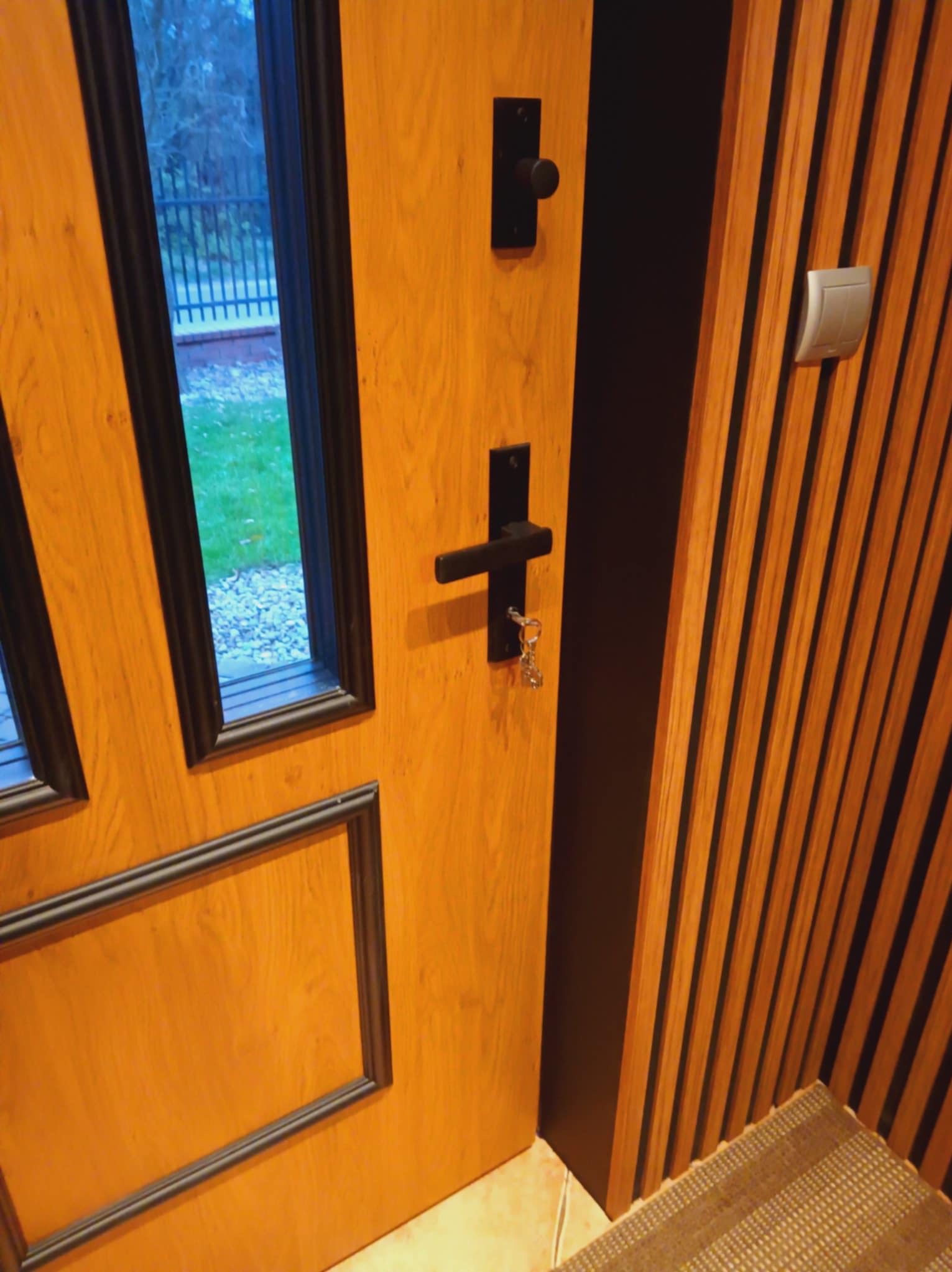 Drewniane drzwi wejściowe z czarnymi detalami i widokiem na zewnątrz, sąsiadujące z ścianą wykończoną pionowymi lamelami w ciepłym odcieniu drewna.