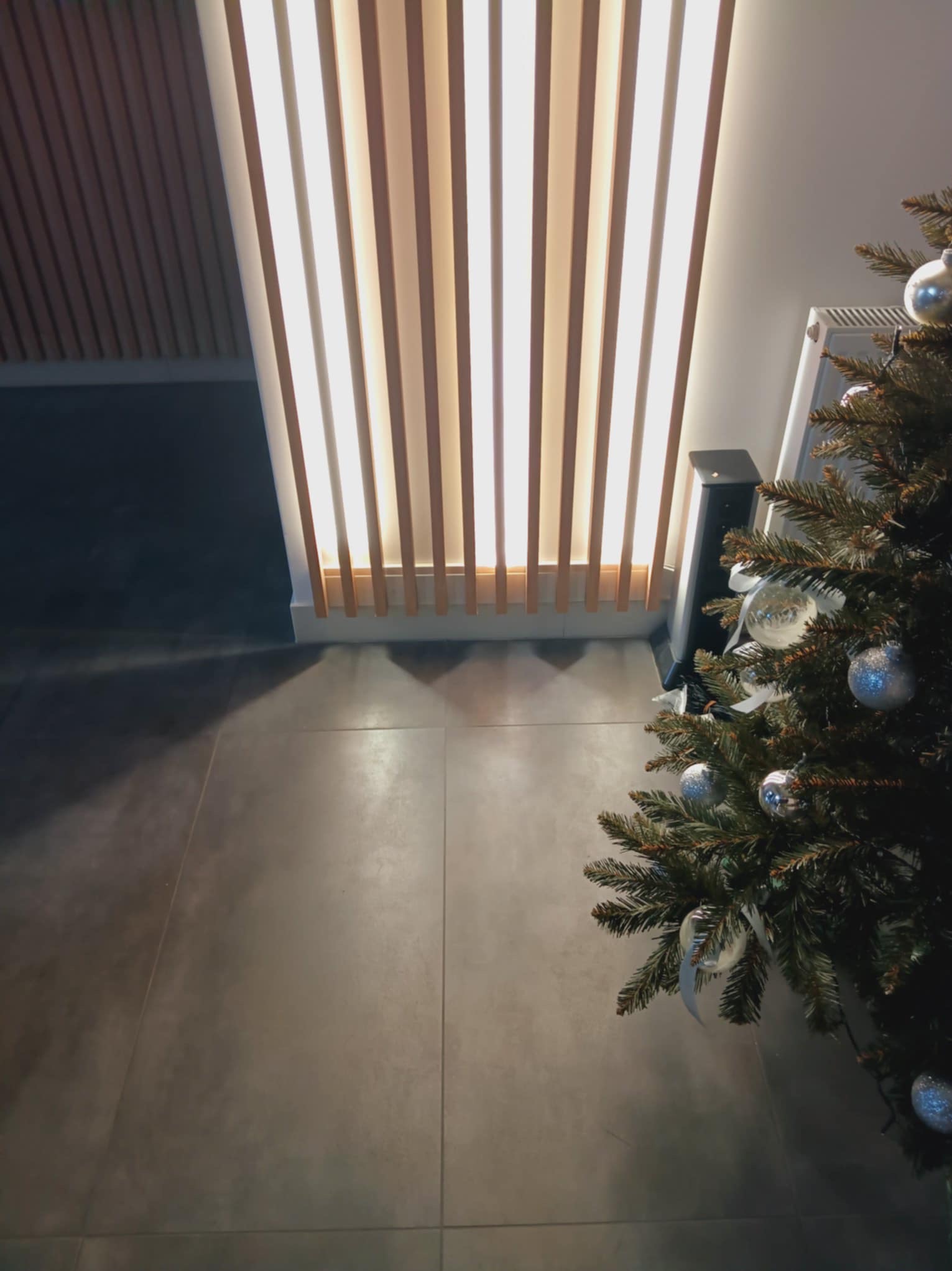 Nowoczesna aranżacja wnętrza z podświetlaną ścianą z drewnianych paneli, szare płytki podłogowe i fragment choinki z srebrnymi bombkami w rogu.