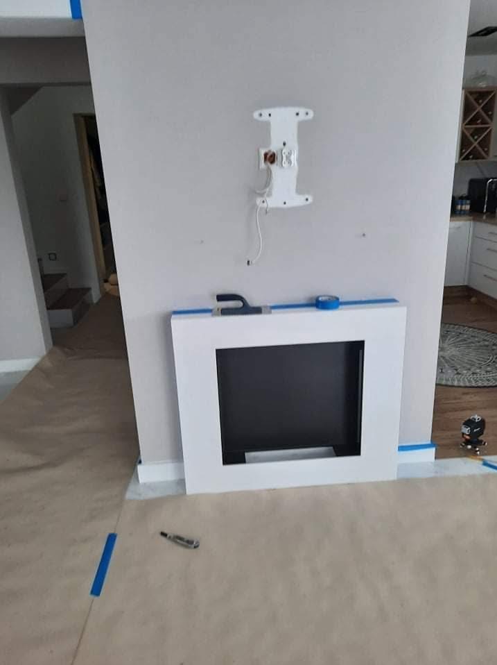 Biała obudowa kominka z czarnym wkładem, zabezpieczona taśmą malarską, na podłodze papier ochronny. Przygotowania do montażu telewizora na ścianie.