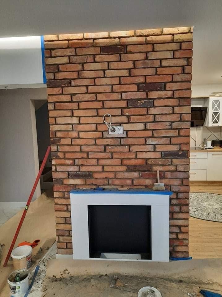 Ściana z cegły klinkierowej z atrapą kominka w trakcie wykańczania. Widoczne narzędzia i zabezpieczenia. W tle fragment kuchni.