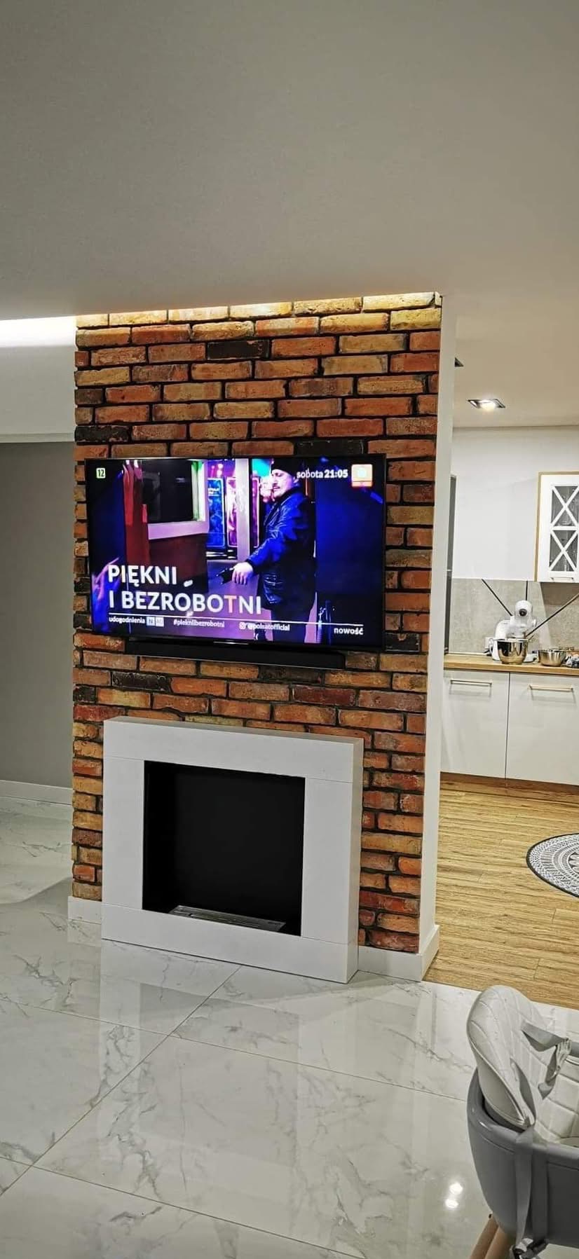Ściana z cegły z telewizorem i kominkiem w nowoczesnym salonie z błyszczącą podłogą. Widok na kuchnię w tle. Oświetlenie LED podkreśla strukturę cegły.