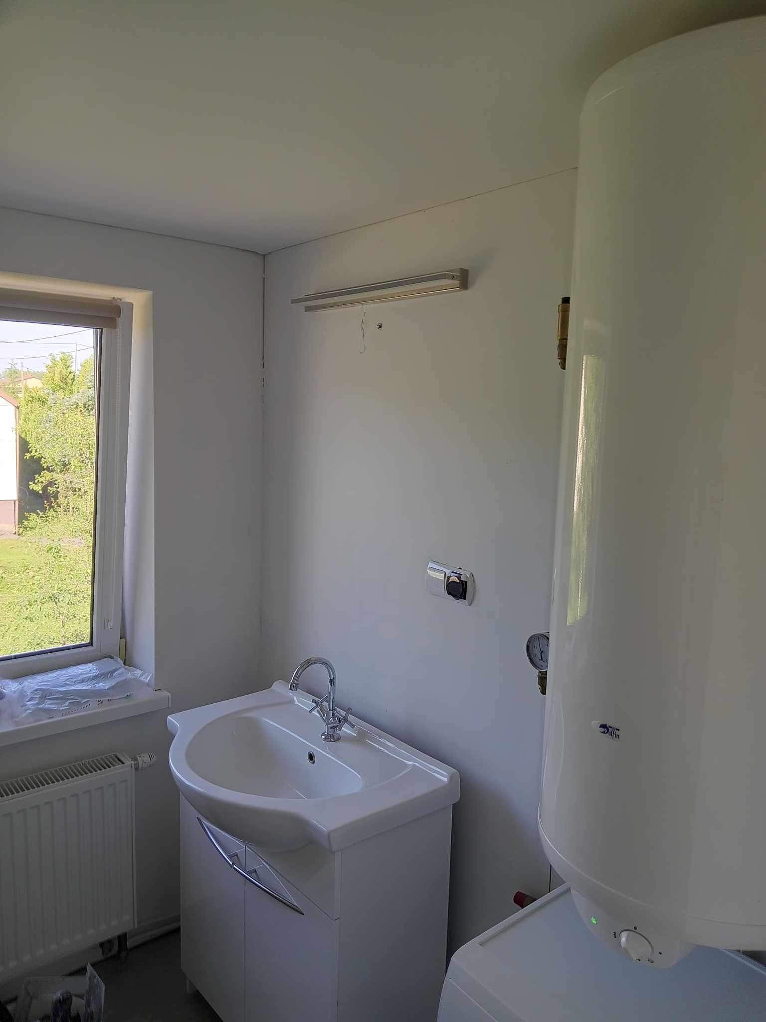 Jasna łazienka z białą umywalką na szafce, grzejnikiem i bojlerem. Okno z widokiem na zieleń. Proste oświetlenie nad umywalką. Nowoczesne wykończenie.