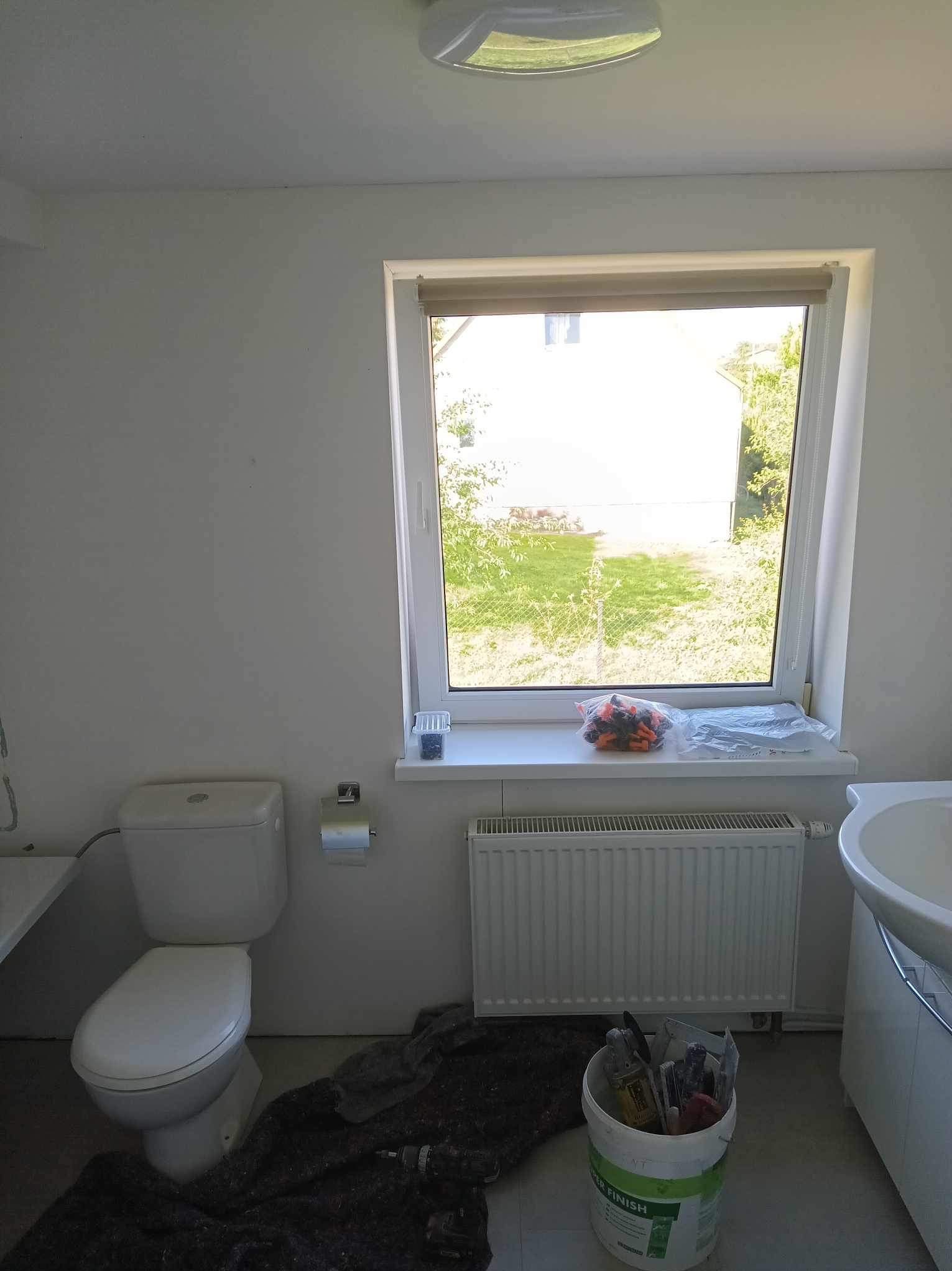 Biała łazienka w trakcie remontu: toaleta, grzejnik, okno z widokiem na zieleń, narzędzia w wiadrze, rozłożona folia ochronna na podłodze.