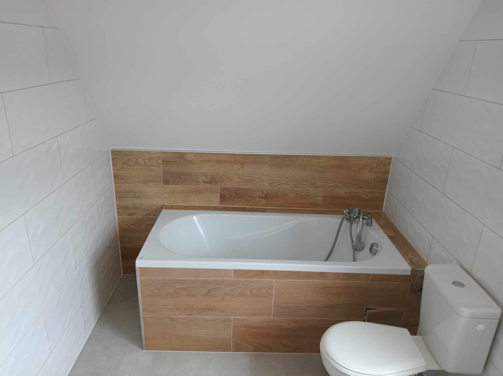 Nowoczesna łazienka na poddaszu z wanną obudowaną płytkami imitującymi drewno, białe płytki na ścianach i szara podłoga. Ukośny sufit.