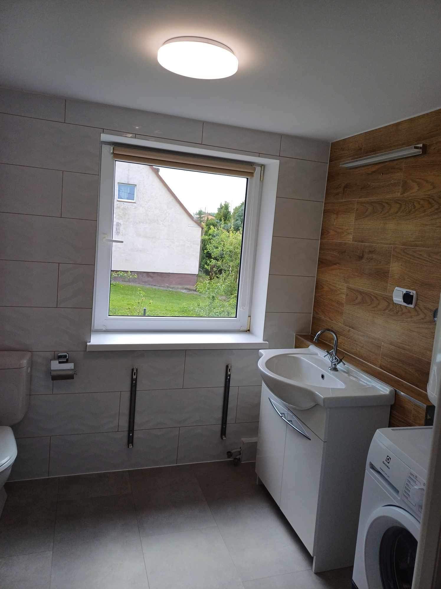 Jasna łazienka z oknem na ogród: płytki imitujące kamień, drewnopodobna ściana, biała umywalka z szafką i pralka. Nowoczesne oświetlenie sufitowe.