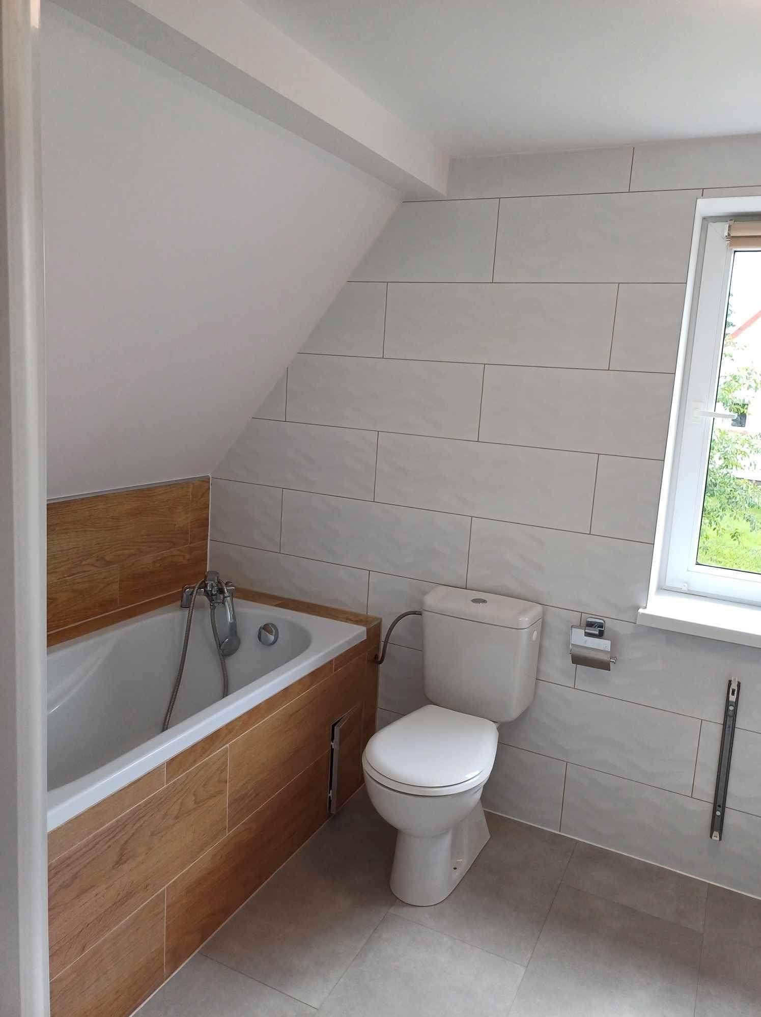 Jasna łazienka na poddaszu z wanną obudowaną drewnopodobnymi płytkami, toaletą i oknem. Ściany wyłożone płytkami w odcieniach szarości. Ukończony remont.