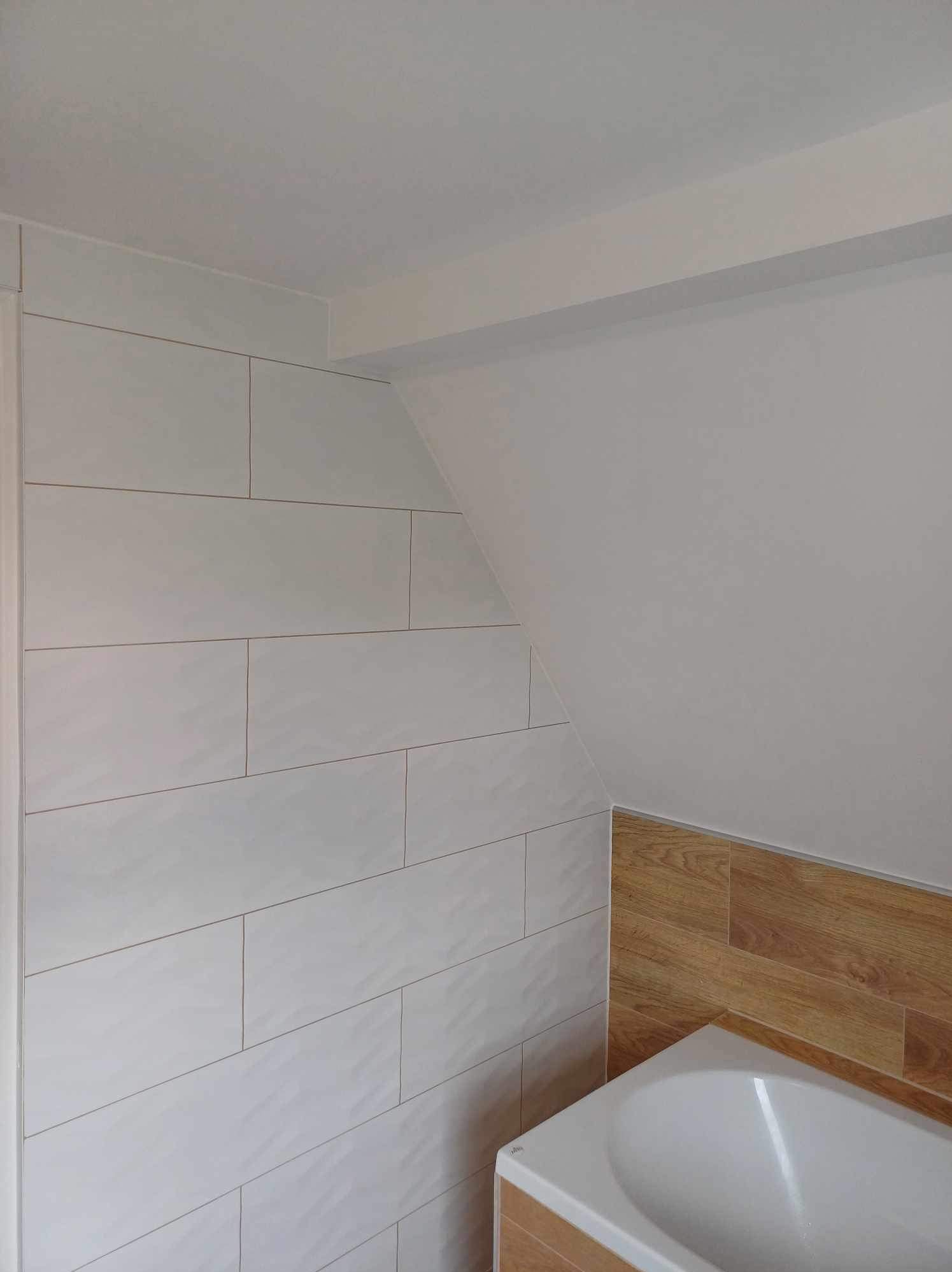 Białe, podłużne płytki w łazience na poddaszu, z drewnopodobnym akcentem przy wannie. Ukośny sufit, minimalistyczny design, jasne wnętrze.
