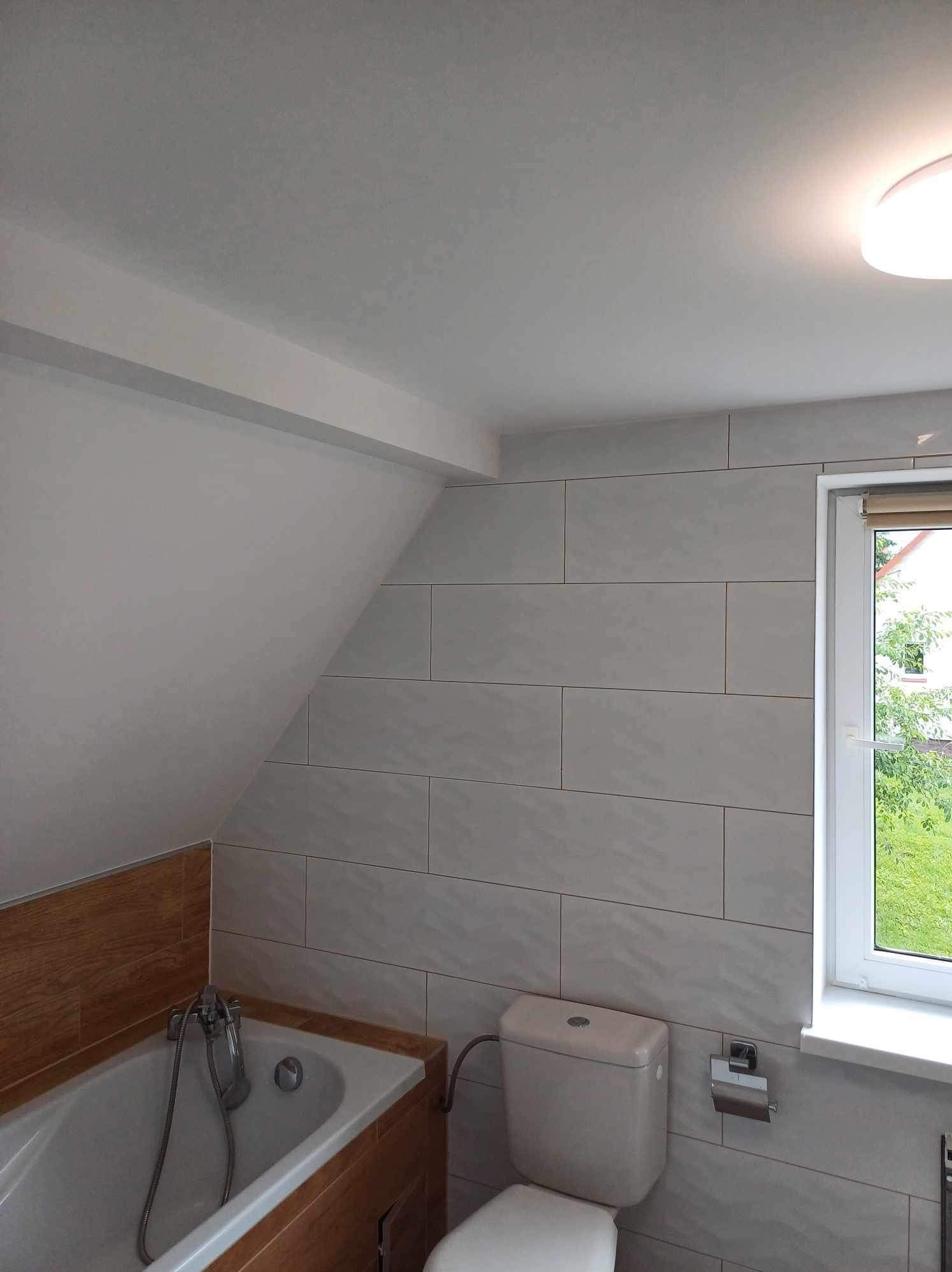 Jasna łazienka na poddaszu z wanną obudowaną drewnem i toaletą. Ściany wyłożone płytkami w odcieniach bieli. Okno z widokiem na zieleń.