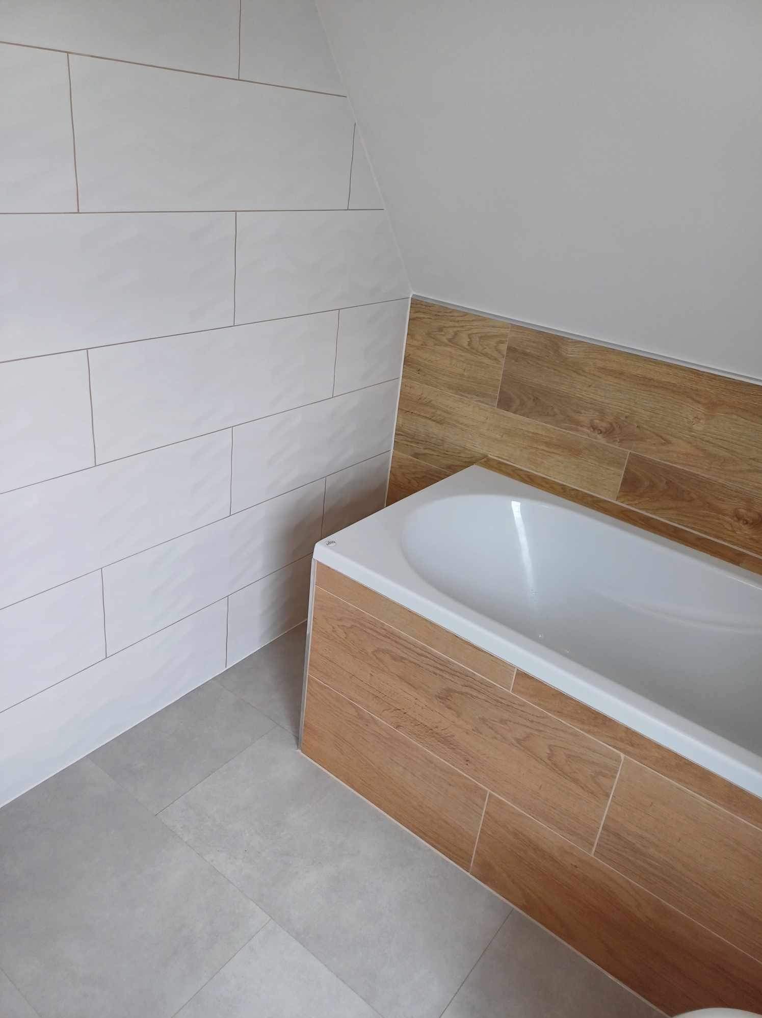 Wykończona łazienka na poddaszu: białe płytki ścienne, drewnopodobna obudowa wanny, szare płytki podłogowe. Ukośny sufit.