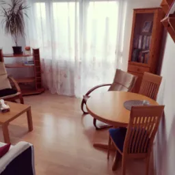 Jasny pokój z drewnianymi meblami: stół z krzesłami, regał z książkami, fotel. Na stole serweta i szklanka. W tle okno z delikatnymi zasłonami i roślina w doniczce.