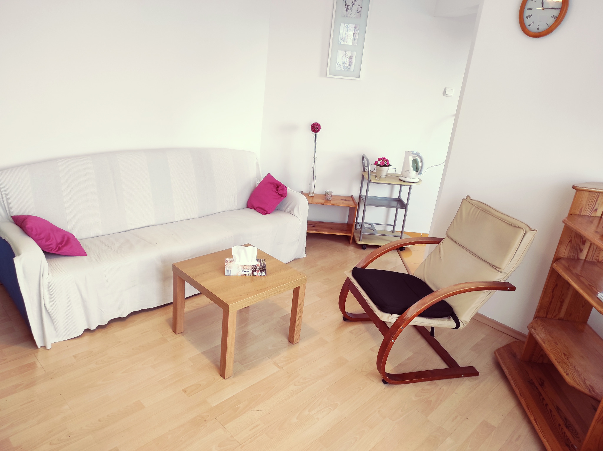 Jasne wnętrze gabinetu z kanapą, fotelem i stolikiem. Na ścianie zegar i grafika. Stonowana kolorystyka sprzyja relaksacji.