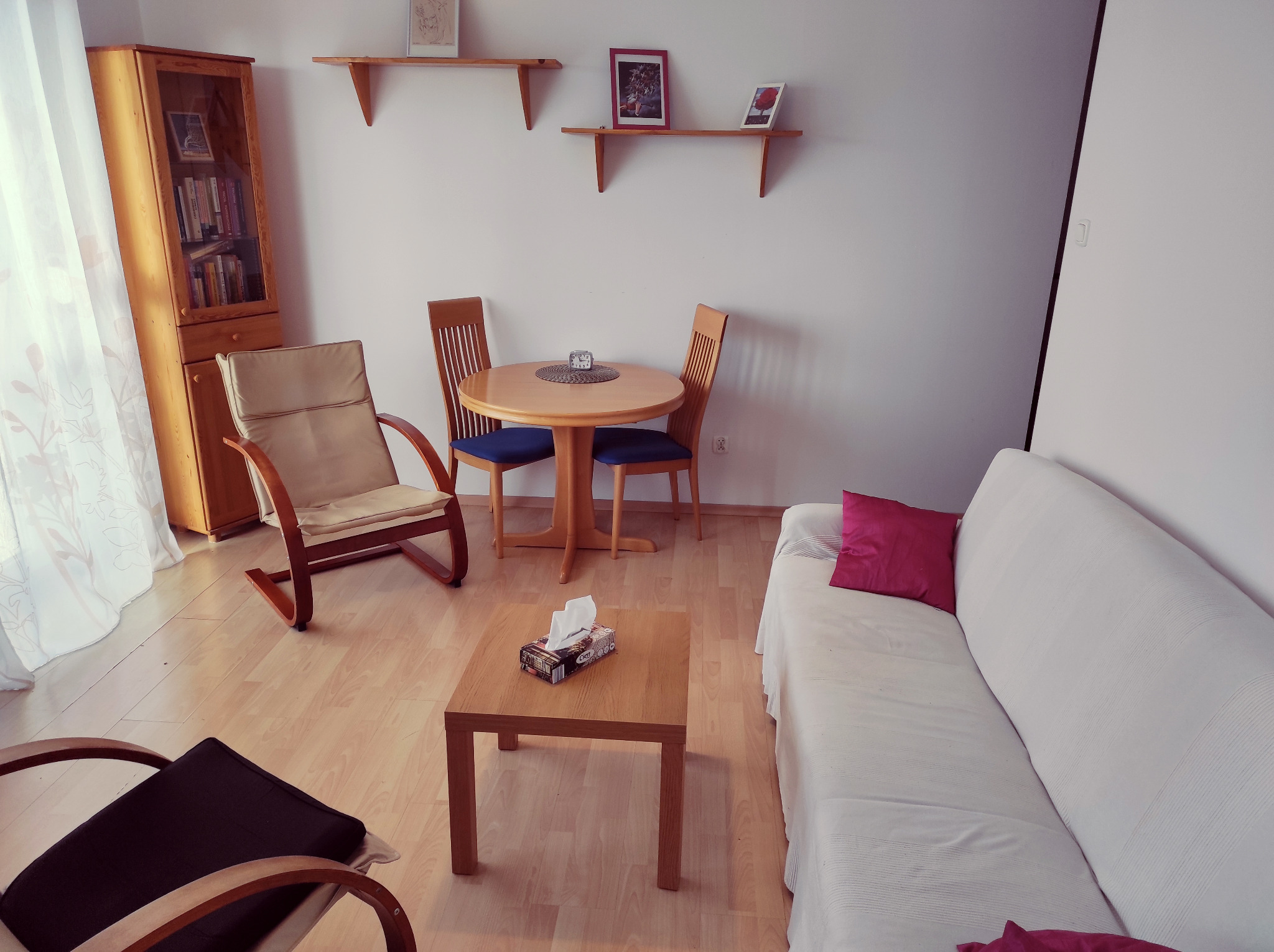 Jasny pokój dzienny z drewnianymi meblami: stół, krzesła, regał, kanapa z poduszką, tworząc przytulną przestrzeń do rozmowy i relaksu.