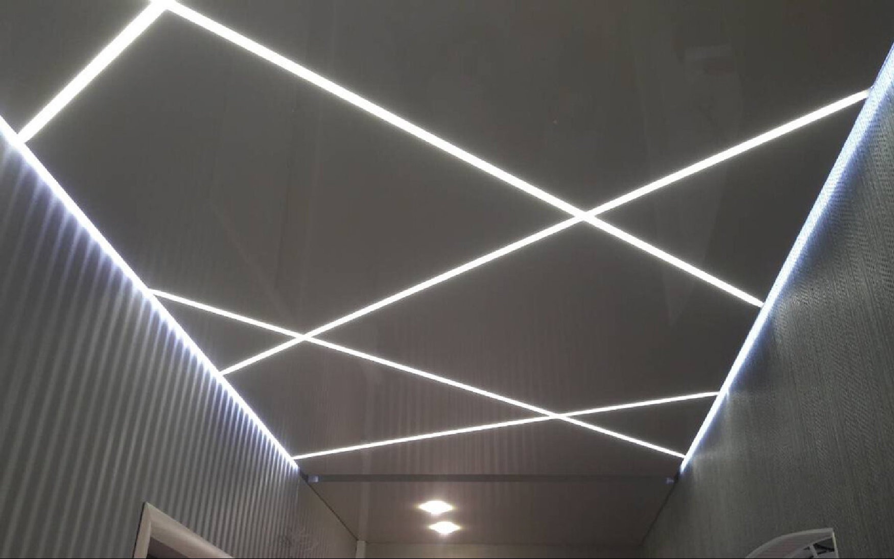 Sufit napinany z geometrycznym układem oświetlenia LED wzdłuż ścian i krzyżujących się linii na powierzchni, tworzący nowoczesny design.