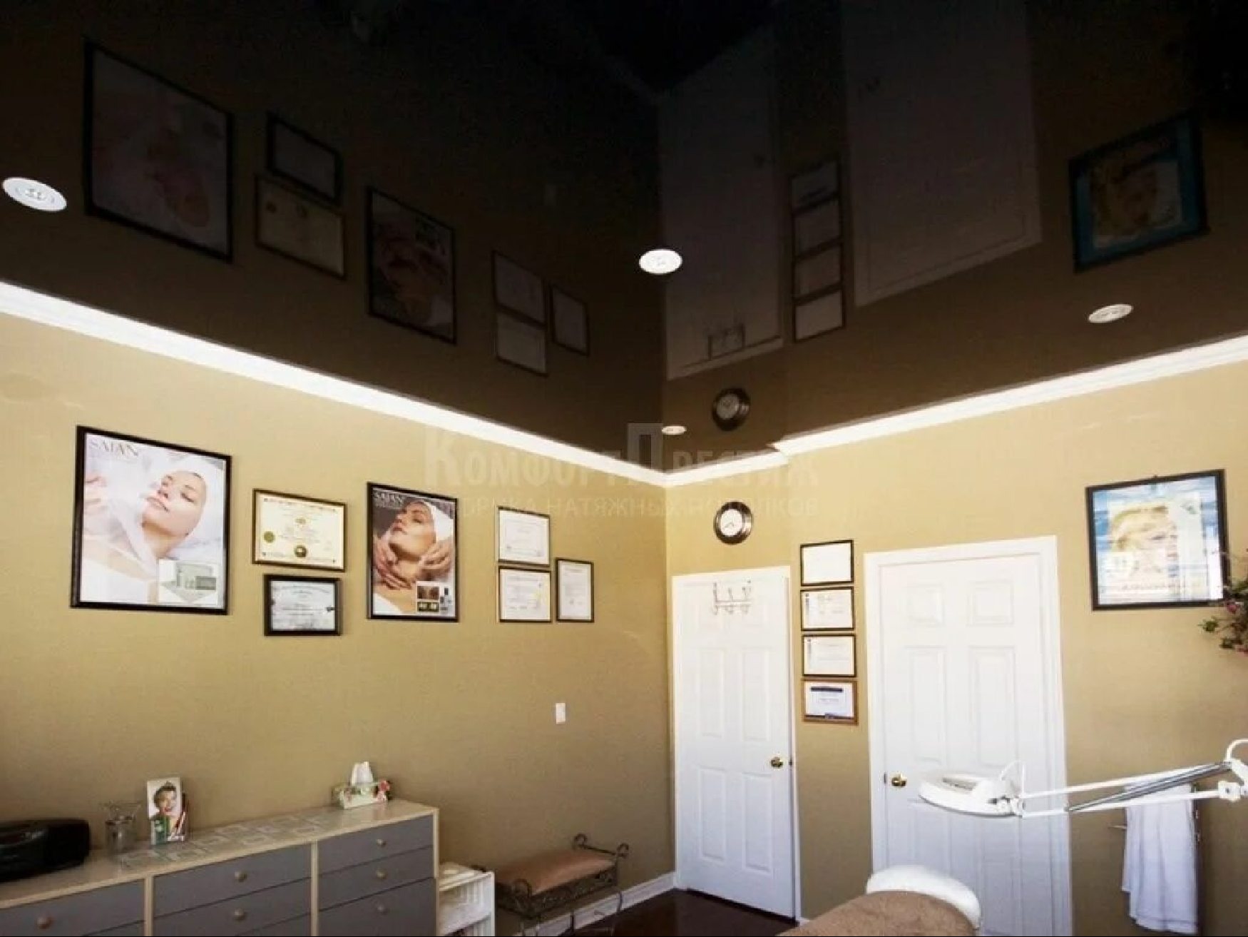 Ciemnobrązowy, błyszczący sufit napinany z oświetleniem punktowym w salonie kosmetycznym. Kremowe ściany zdobią obrazy i certyfikaty.