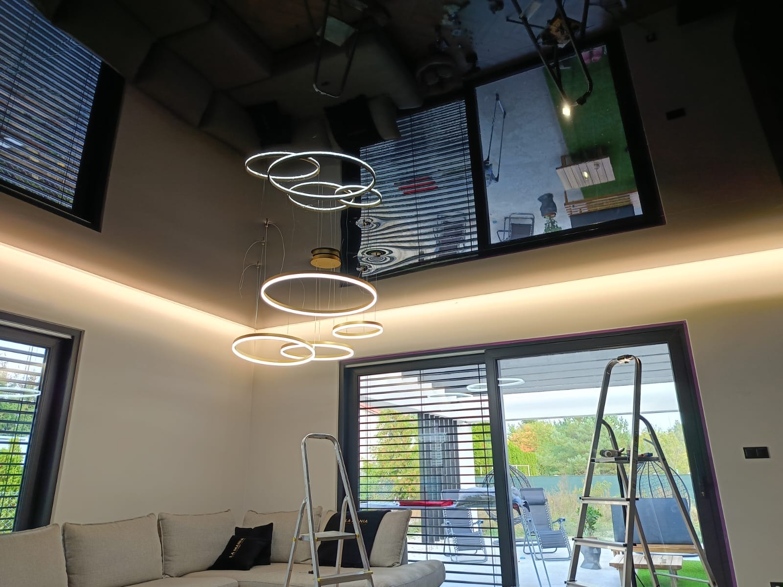 Błyszczący, czarny sufit napinany z odbiciem wnętrza, oświetlony ledowym paskiem i nowoczesną lampą pierścieniową. Dwie drabiny stoją w salonie.