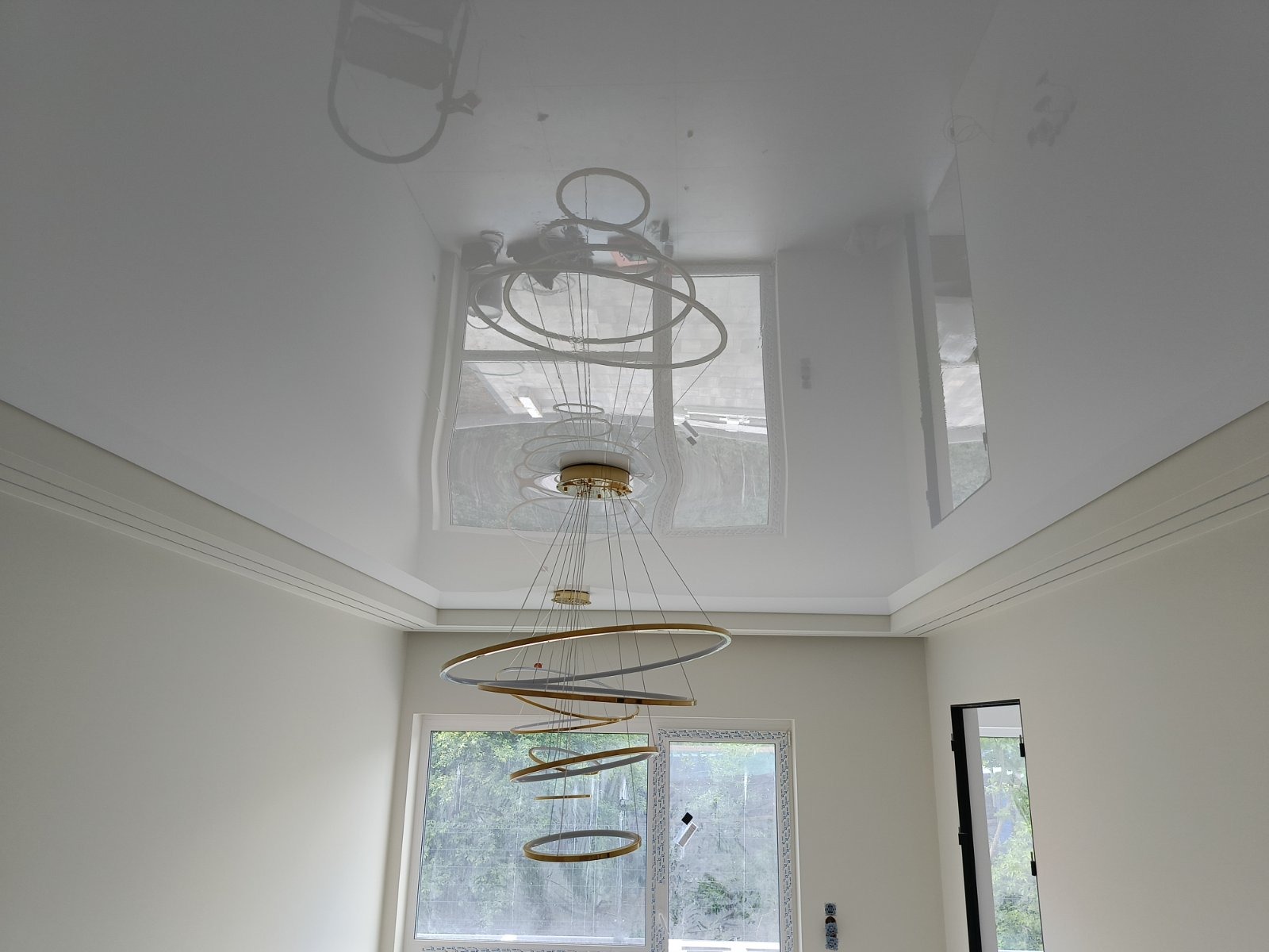 Biały, błyszczący sufit z widocznym odbiciem żyrandola w kształcie okręgów. Nowoczesny design wnętrza z oknem i listwami dekoracyjnymi.
