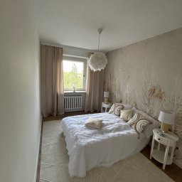 TOL-BUD Anatolii Staryk - Jasna sypialnia z tapetą w pióra, miękkim dywanem i białym łóżkiem. Okno z beżowymi zasłonami. Dwie białe szafki nocne z lampkami.