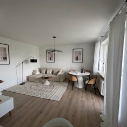 TOL-BUD Anatolii Staryk - Jasne, przestronne wnętrze z drewnianą podłogą, beżową sofą narożną, okrągłym stołem i białymi ścianami z cegły. Minimalistyczny wystrój, lampa podłogowa i abstrakcyjne obrazy.