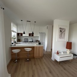 TOL-BUD Anatolii Staryk - Jasne wnętrze: kuchnia z wyspą i hokerami, drewniana podłoga, minimalistyczny design, białe ściany, lampy wiszące, dekoracje. Widok z perspektywy wejścia.