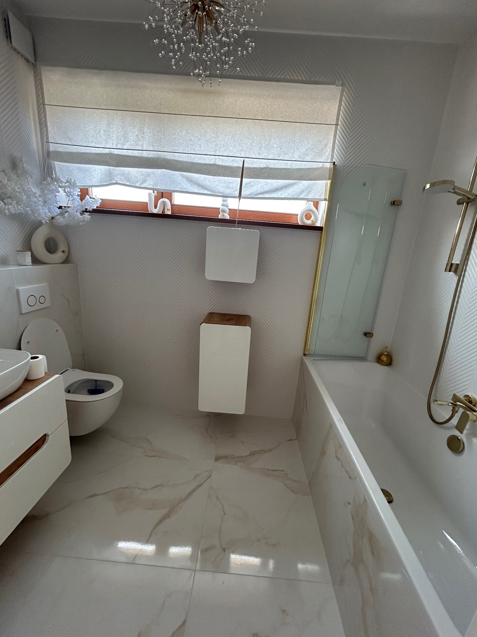 Elegancka, jasna łazienka po remoncie z marmurową podłogą, wiszącą toaletą i złotymi akcentami armatury. W oknie roleta rzymska i dekoracje.