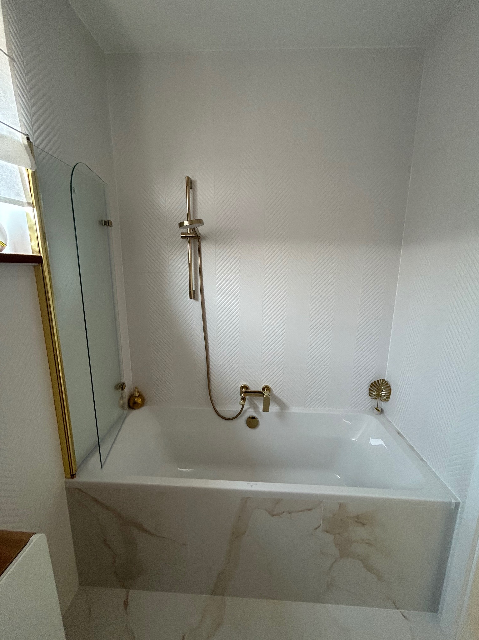 Elegancka, biała łazienka z wanną obudowaną marmurem, złotą armaturą i dekoracyjną ścianą w subtelny wzór. Nowoczesny design z dbałością o detale.