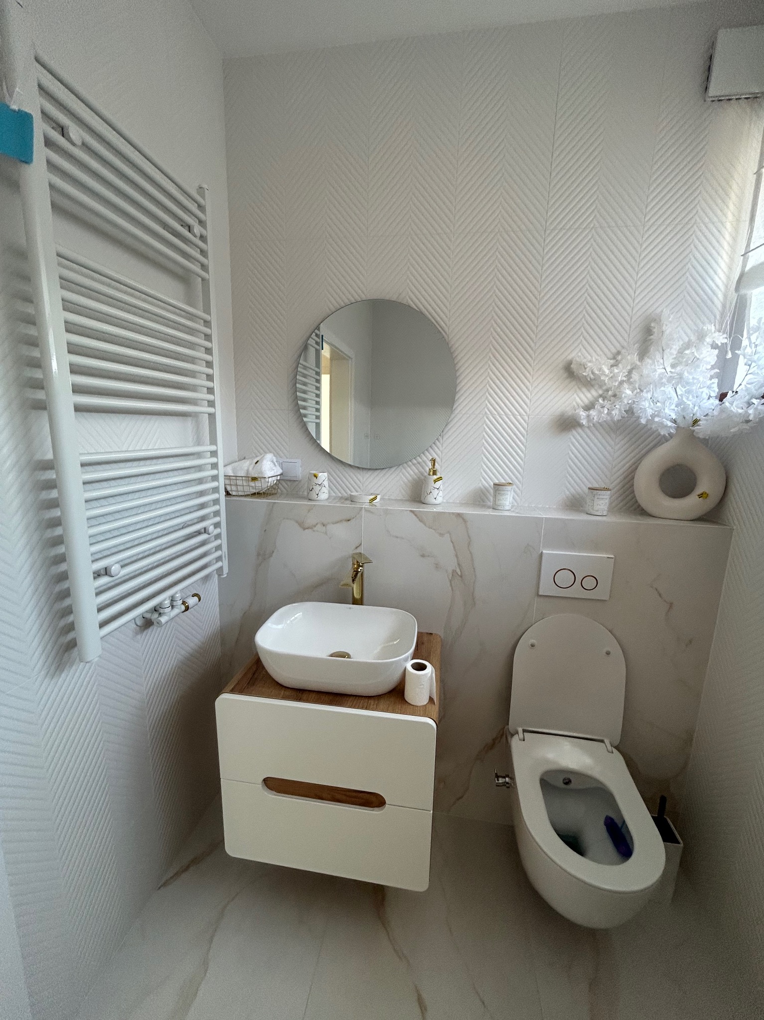 Minimalistyczna, jasna łazienka z wzorzystą tapetą, okrągłym lustrem, umywalką na szafce z drewnianym blatem i grzejnikiem drabinkowym.