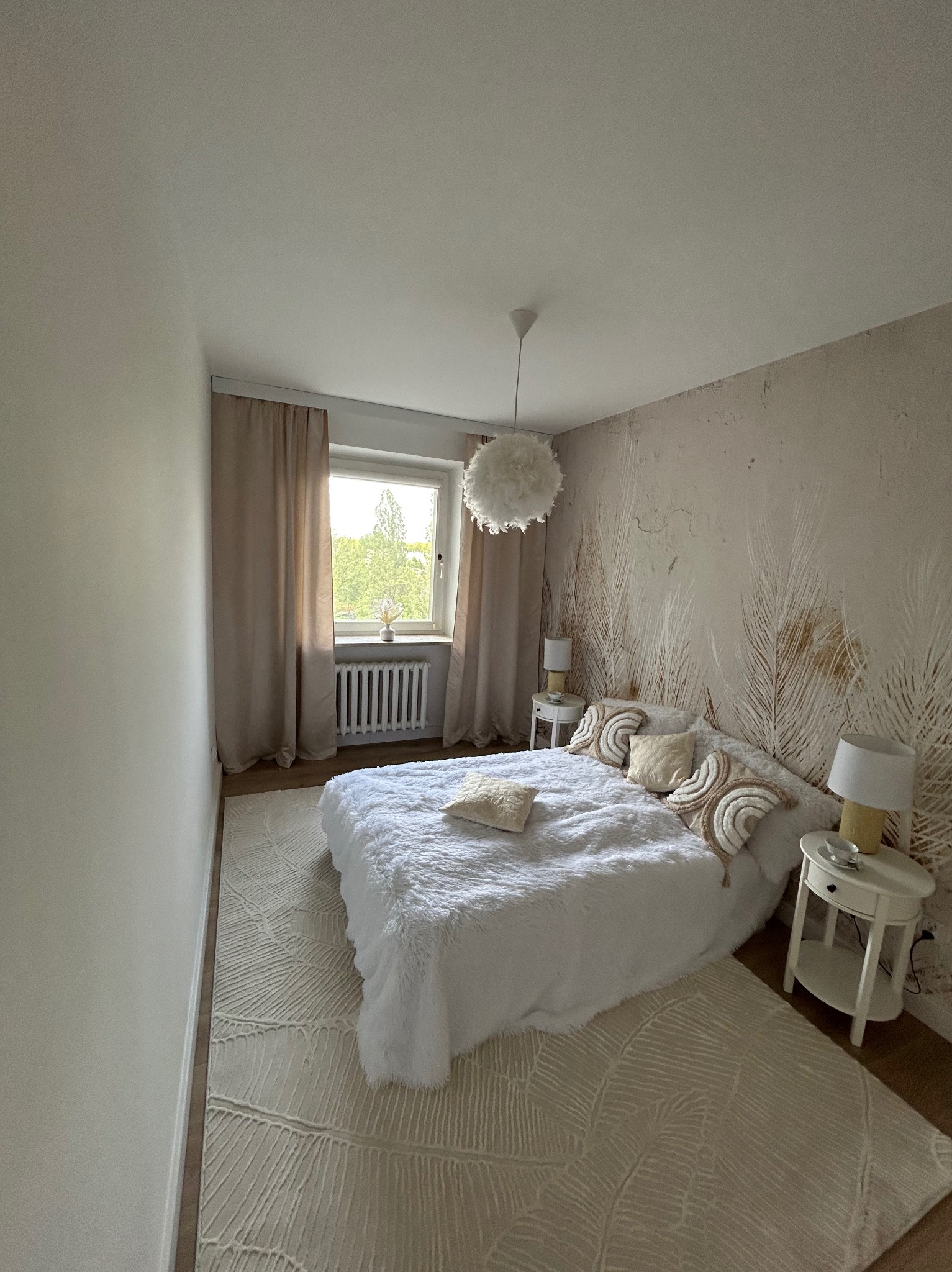 Jasna sypialnia z tapetą w pióra, miękkim dywanem i białym łóżkiem. Okno z beżowymi zasłonami. Dwie białe szafki nocne z lampkami.