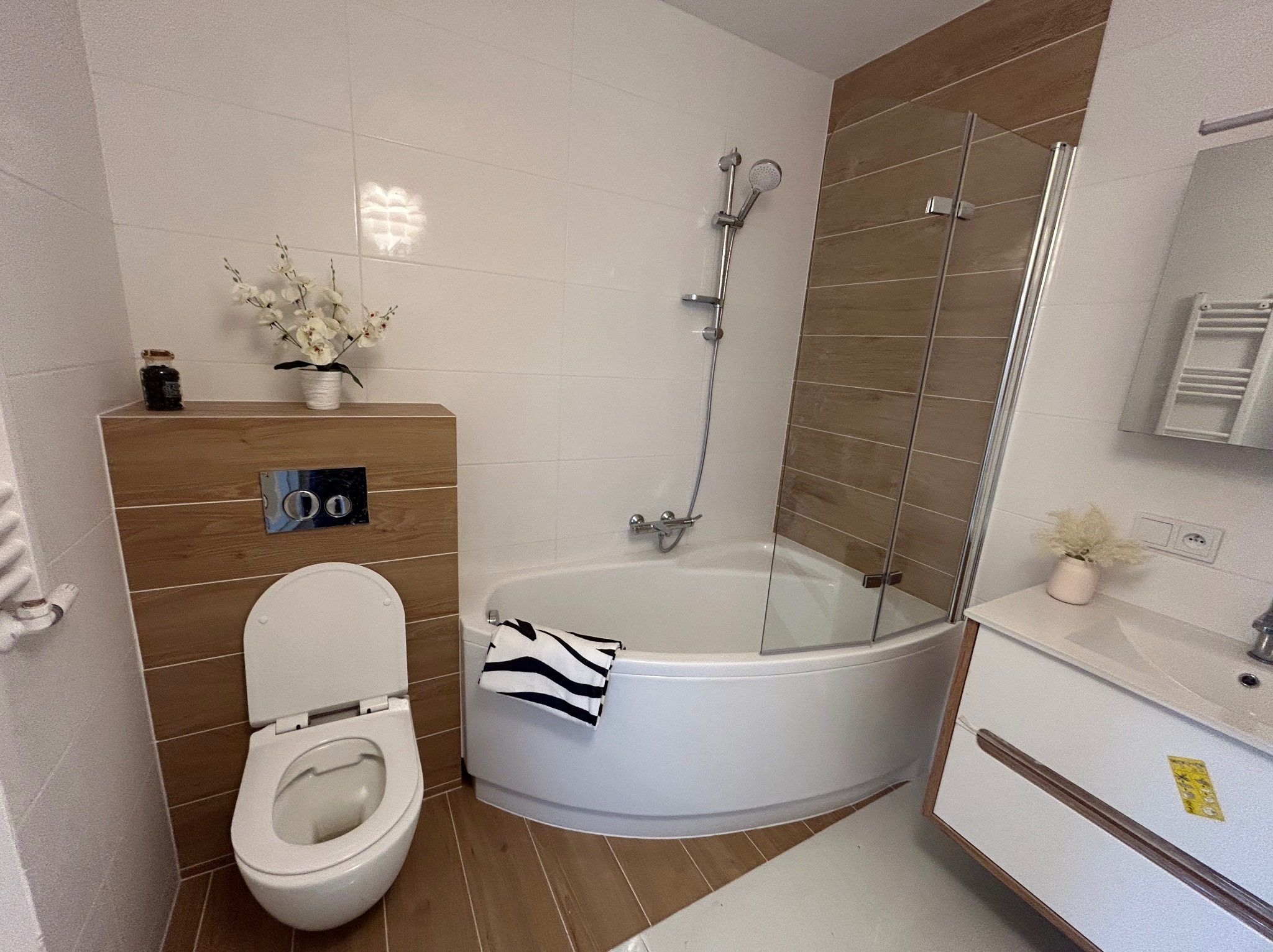 Nowoczesna łazienka po remoncie: białe płytki, drewnopodobne akcenty, narożna wanna z kabiną, toaleta w zabudowie i umywalka z szafką. Całość utrzymana w jasnej kolorystyce.