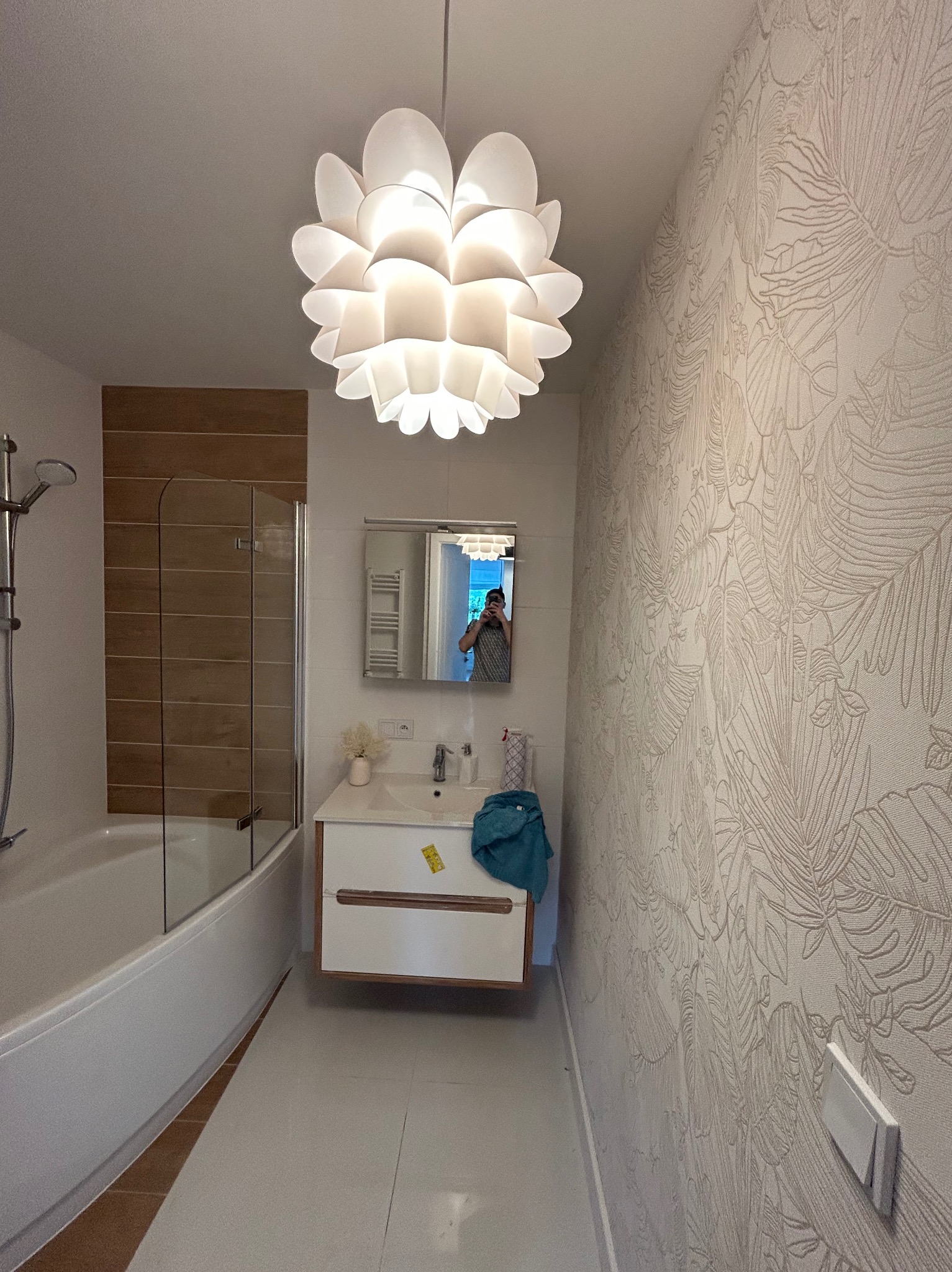 Wykończona łazienka z wanną, umywalką i wzorzystą tapetą. Lampa w kształcie kwiatu nadaje wnętrzu nowoczesny charakter. Widoczne detale wykończenia.