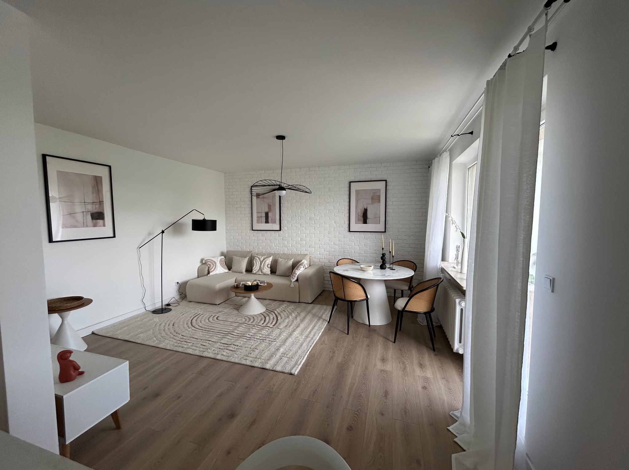 Jasne, przestronne wnętrze z drewnianą podłogą, beżową sofą narożną, okrągłym stołem i białymi ścianami z cegły. Minimalistyczny wystrój, lampa podłogowa i abstrakcyjne obrazy.