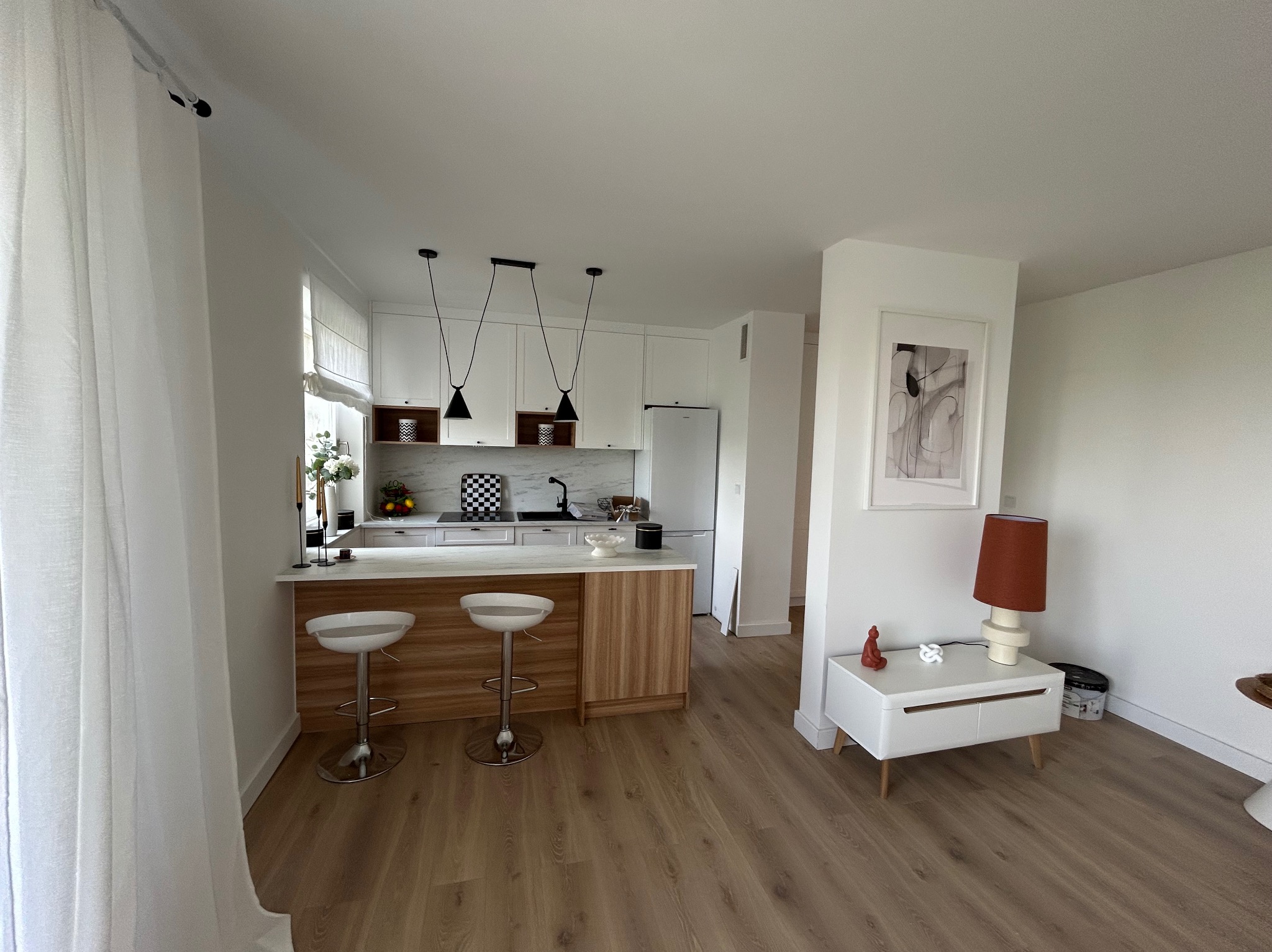 Jasne wnętrze: kuchnia z wyspą i hokerami, drewniana podłoga, minimalistyczny design, białe ściany, lampy wiszące, dekoracje. Widok z perspektywy wejścia.