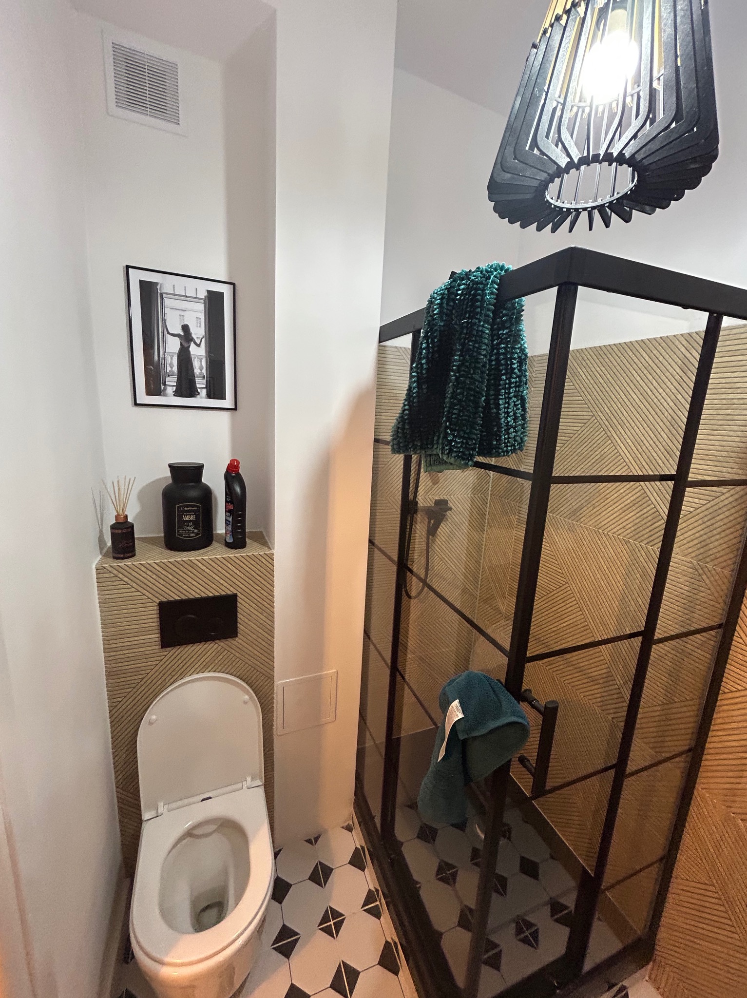 Nowoczesna łazienka z kabiną prysznicową typu walk-in, czarną armaturą i geometrycznymi płytkami podłogowymi. Nad toaletą dekoracje i czarno-biała fotografia.