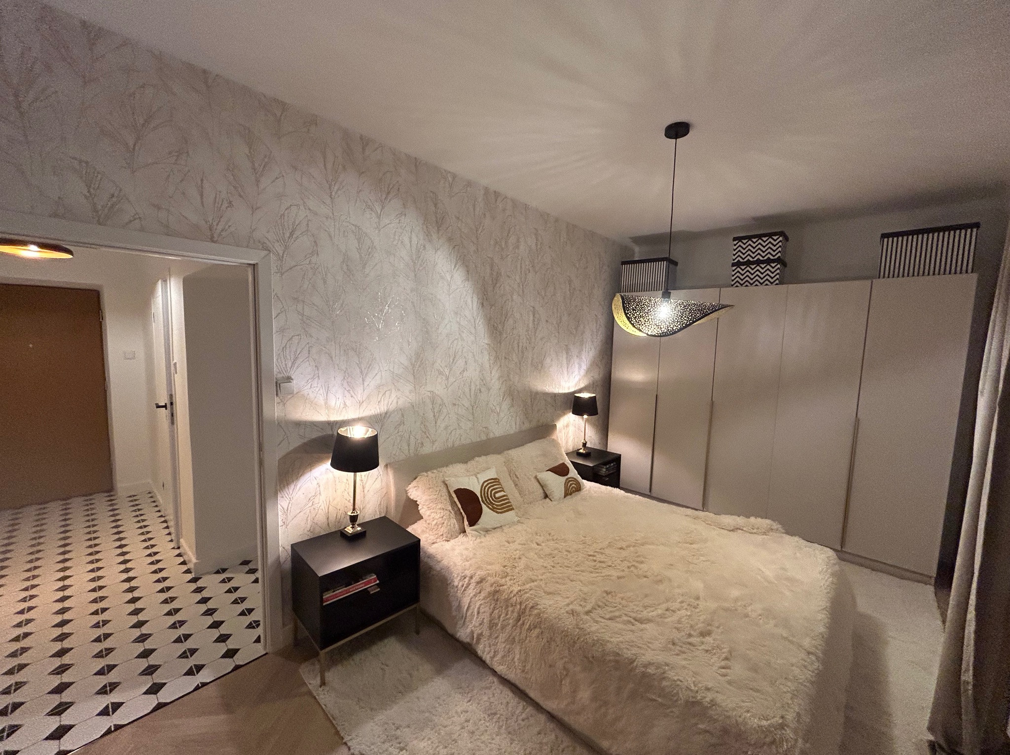 Elegancka sypialnia po remoncie: wzorzysta tapeta, beżowe futrzane łóżko, czarne szafki nocne z lampkami i oryginalna lampa wisząca.