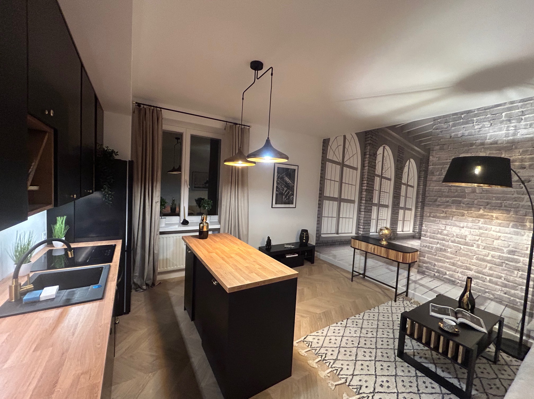 Nowoczesna kuchnia z wyspą, czarne szafki, drewniany blat, designerskie lampy, ceglana fototapeta, parkiet w jodełkę i dywan w geometryczny wzór.