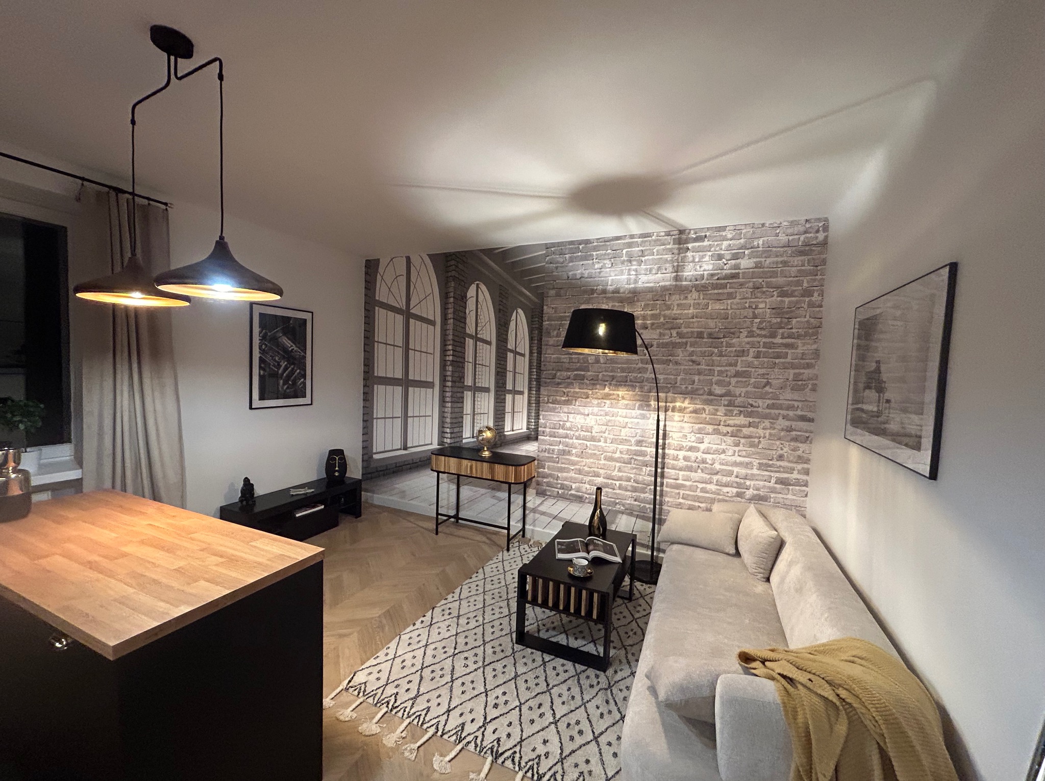 Stylowe wnętrze salonu z ceglaną fototapetą, drewnianym blatem wyspy kuchennej i designerskimi lampami wiszącymi. Uzupełniają je dywan w marokański wzór i nowoczesne meble.