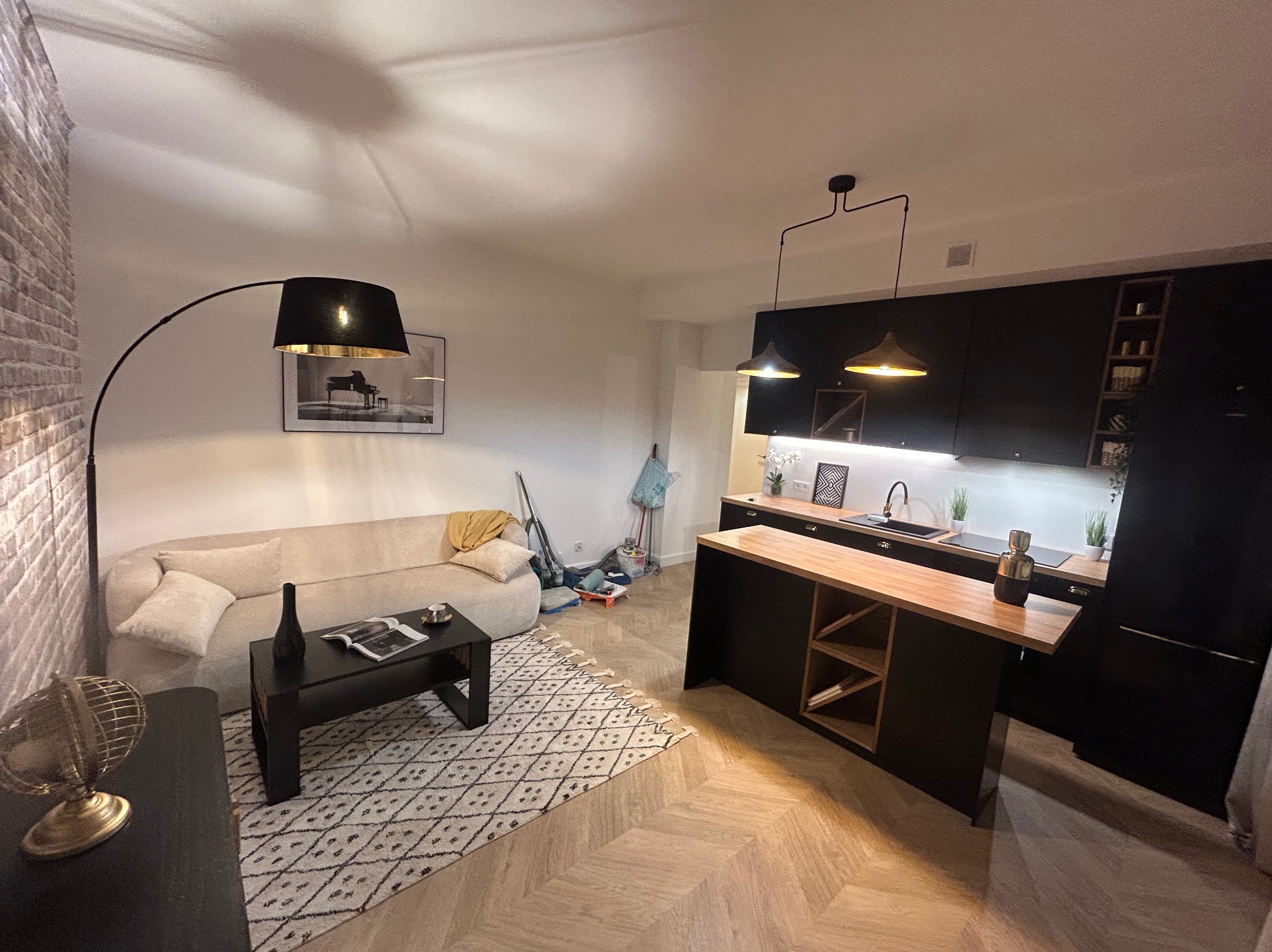 Nowoczesne wnętrze: salon z aneksem kuchennym. Parkiet w jodełkę, czarna zabudowa kuchenna, beżowa sofa, lampa łukowa. Minimalistyczny design z dbałością o detale.