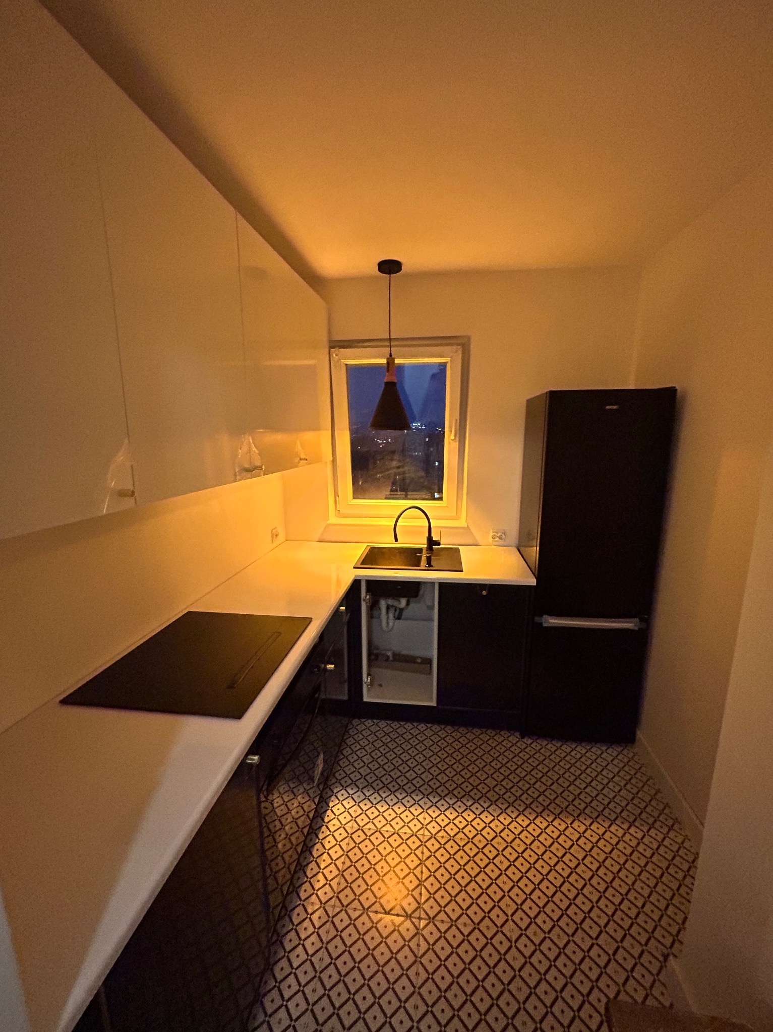 Nowoczesna, mała kuchnia z czarnymi akcentami: szafki, zlew, lodówka. Białe blaty i górne szafki. W tle okno z widokiem na miasto nocą i wzorzysta podłoga.