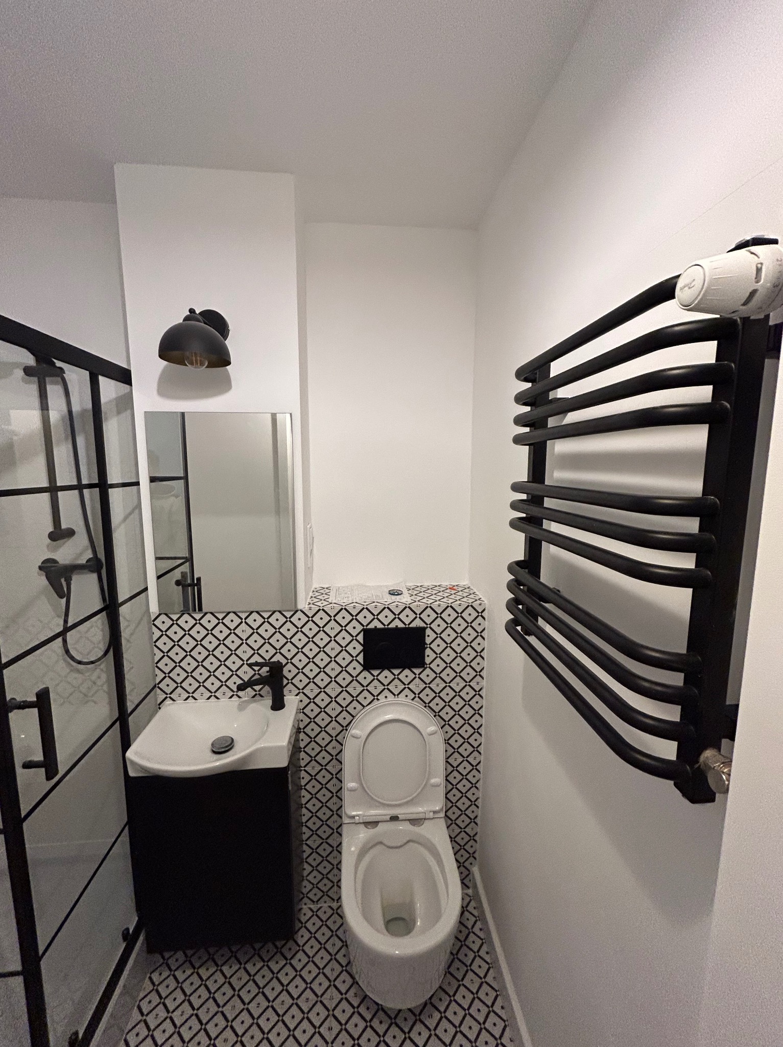 Nowoczesna, czarno-biała łazienka z kabiną prysznicową typu walk-in, umywalką z szafką, toaletą i grzejnikiem drabinkowym. Wzór płytek w romby.