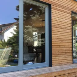 Nowoczesne okna przesuwne z szarymi ramami w drewnianej elewacji domu. Widoczne odbicie otoczenia w szybach. Minimalistyczny design i naturalne materiały.