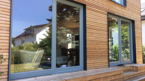 Nowoczesne okna przesuwne z szarymi ramami w drewnianej elewacji domu. Widoczne odbicie otoczenia w szybach. Minimalistyczny design i naturalne materiały.