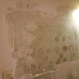 Michał - Ściana w trakcie remontu z widoczną siatką wzmacniającą i częściowo nałożoną gładzią. Widoczny włącznik światła i lampa. Ujęcie z bliska.