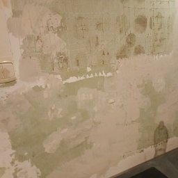 Michał - Przygotowanie ściany do remontu: usunięte stare powłoki, widoczna siatka wzmacniająca, obok włącznik światła i butelka wody. Ściana w trakcie prac przygotowawczych.