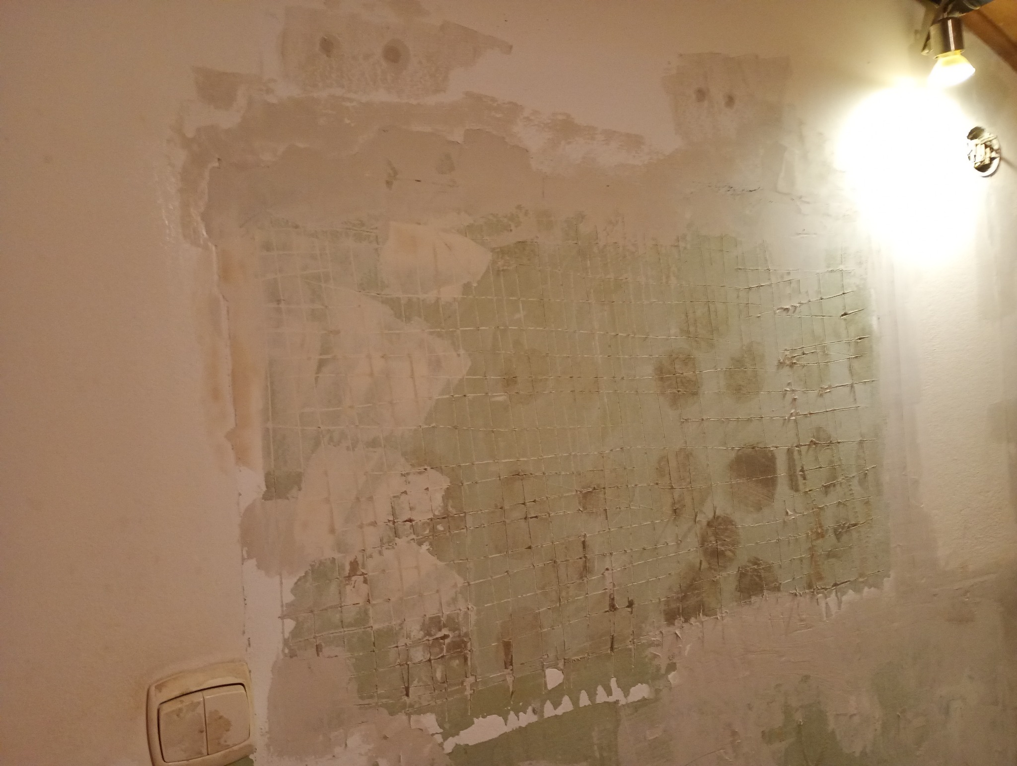 Ściana w trakcie remontu z widoczną siatką wzmacniającą i częściowo nałożoną gładzią. Widoczny włącznik światła i lampa. Ujęcie z bliska.
