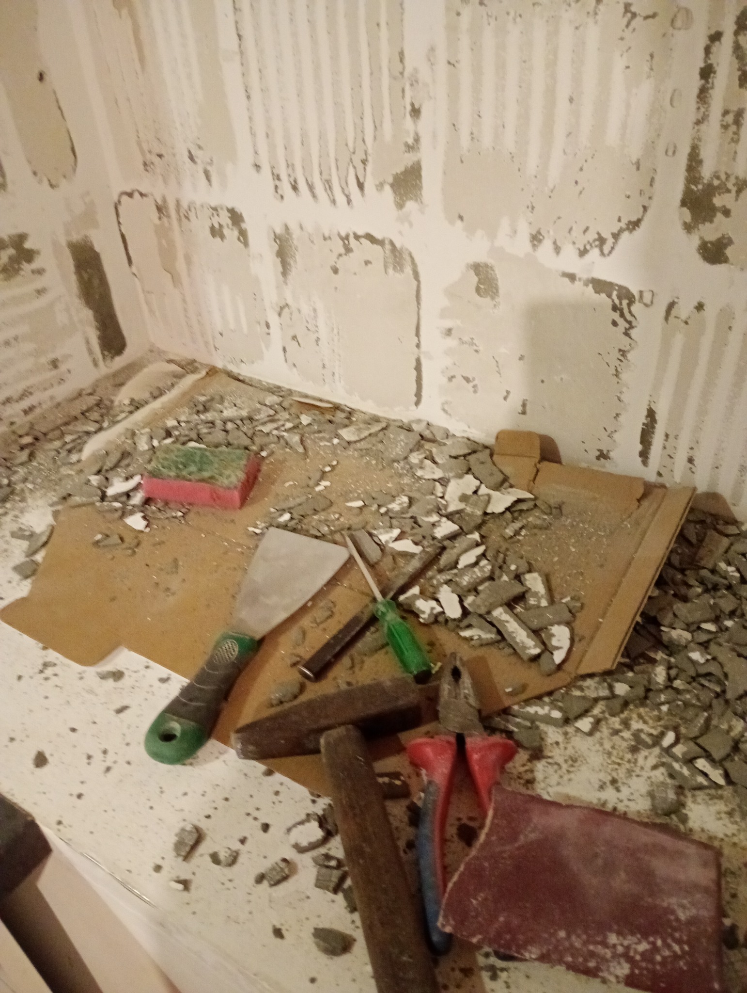 Fragment ściany po skuciu płytek z widocznymi narzędziami: szpachelka, młotek, szczypce. Gruz na podłodze. Etap przygotowania podłoża pod nowe płytki.