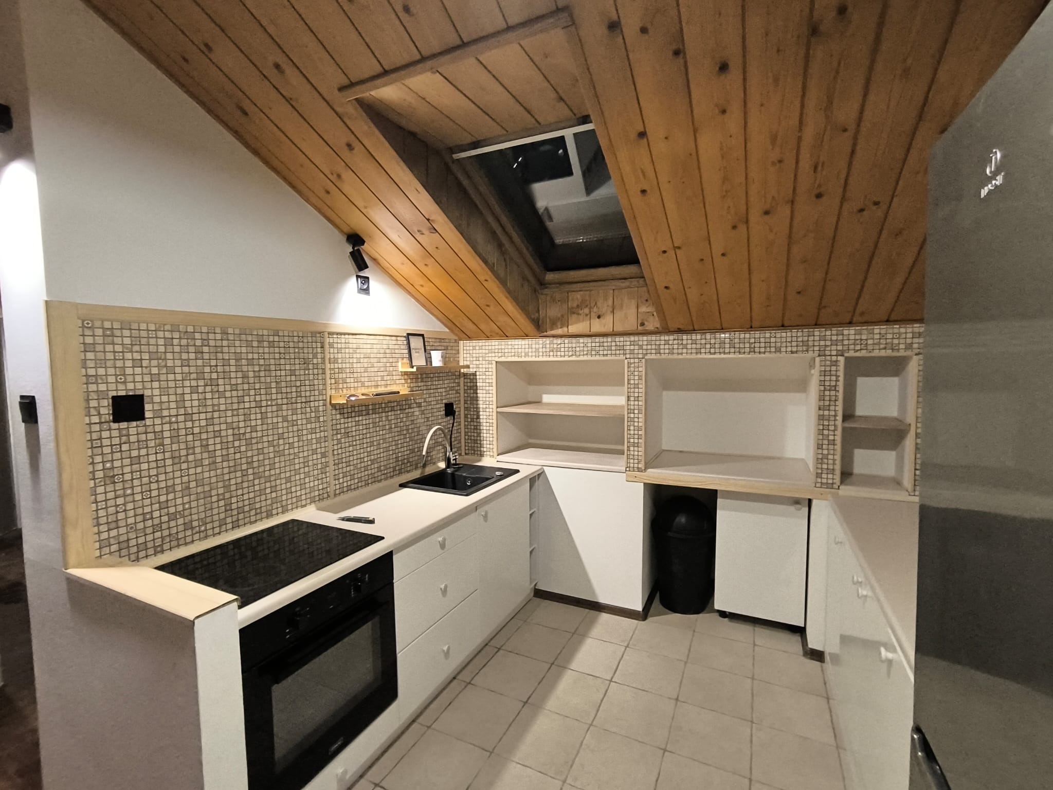 Nowoczesna kuchnia na poddaszu z drewnianym sufitem i oknem dachowym. Białe szafki, mozaika nad blatem, czarny zlew i płyta grzewcza. Widoczny fragment lodówki.