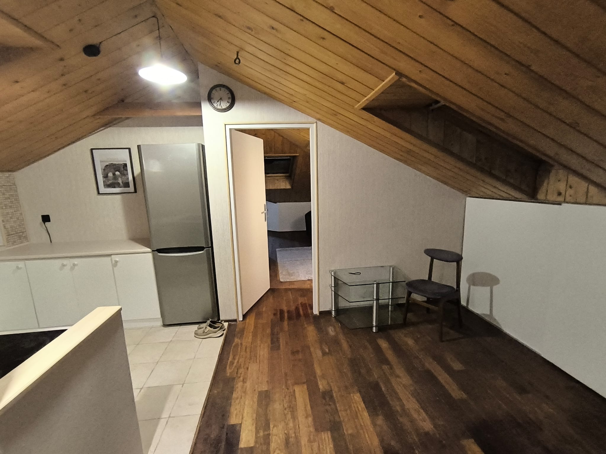 Adaptacja poddasza: drewniana boazeria na skosach, jasna kuchnia z lodówką, widok na pokój przez otwarte drzwi, zegar na ścianie. Buty leżą przy lodówce.