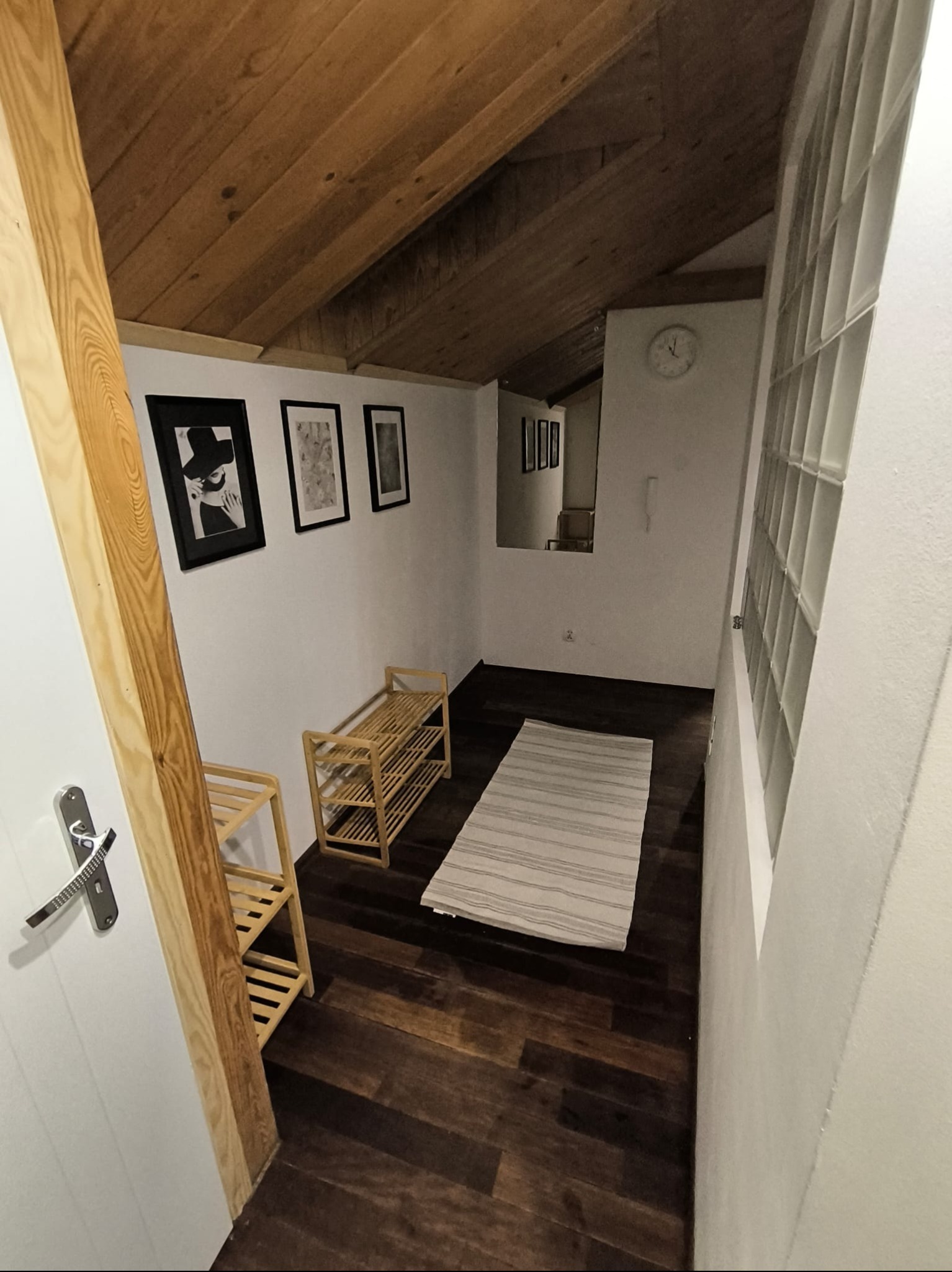 Hall z drewnianą podłogą i panelami na suficie, białe ściany z grafikami w czarnych ramkach, drewniane półki na buty i dywanik w paski. Widok z perspektywy wejścia.