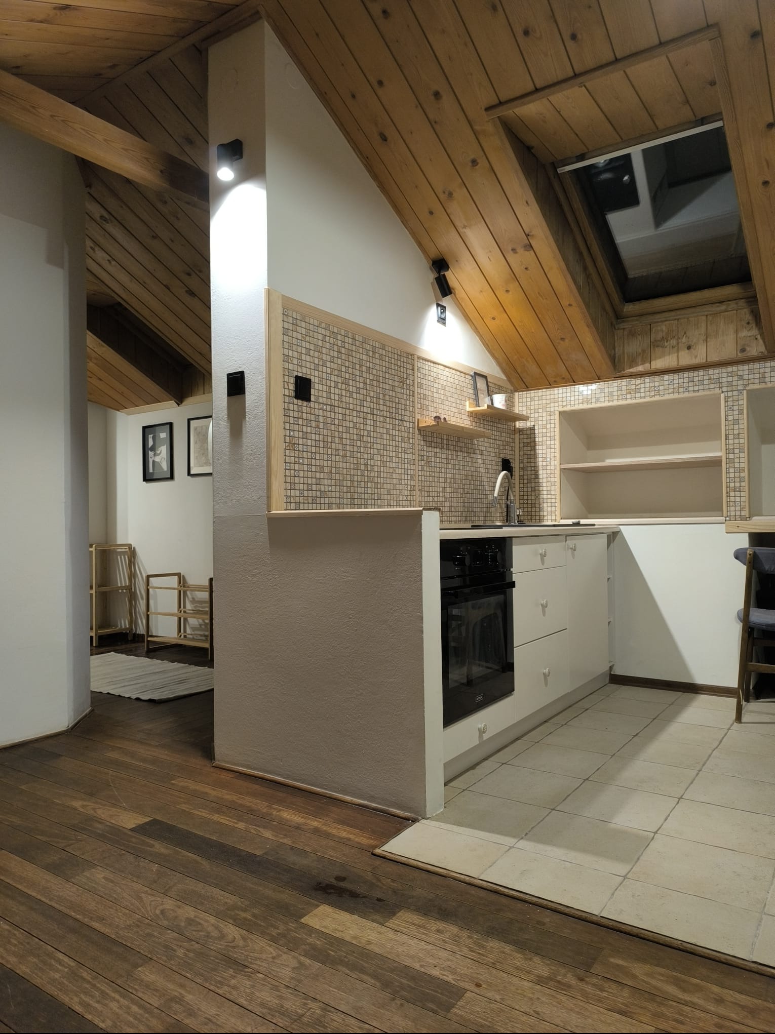 Kuchenne wnętrze z drewnianym sufitem i oknem połaciowym, mozaiką nad blatem i nowoczesną zabudową. Parkiet kontrastuje z jasnymi płytkami podłogowymi.