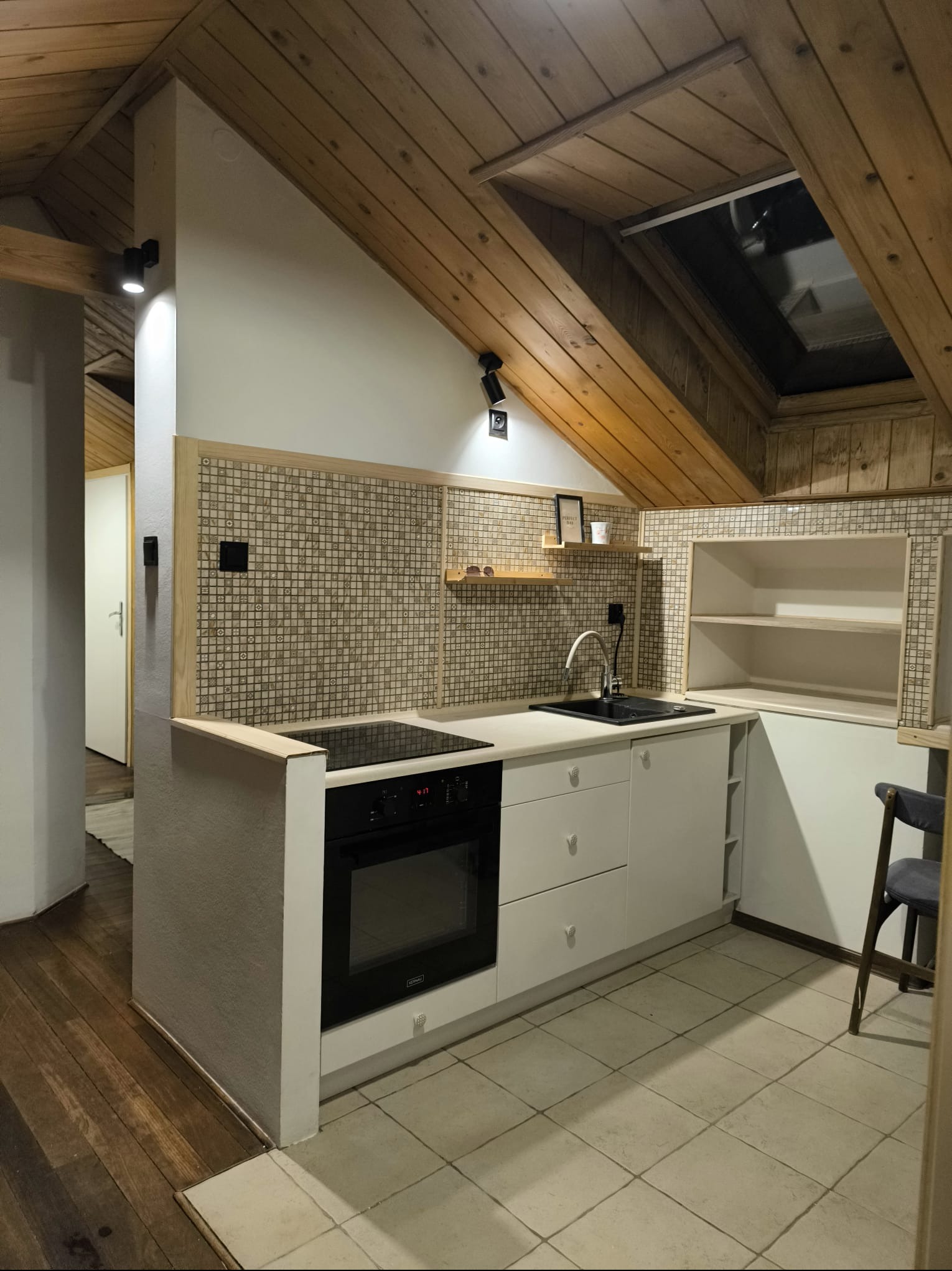 Funkcjonalna kuchnia na poddaszu z skośnym drewnianym sufitem i oknem dachowym. Białe meble, mozaikowa ściana nad blatem i drewniane akcenty.