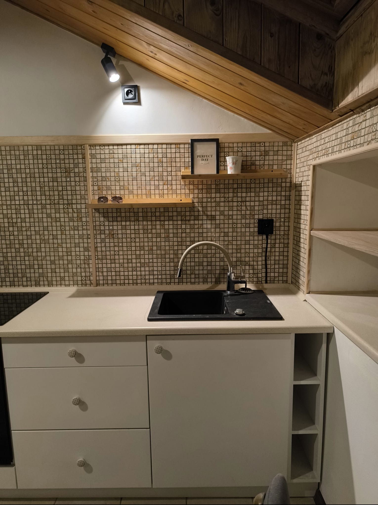Kuchnia na poddaszu: kremowe szafki z mozaikowymi gałkami, czarny zlew, drewniane półki z dekoracjami, mozaika na ścianie i drewniany strop.